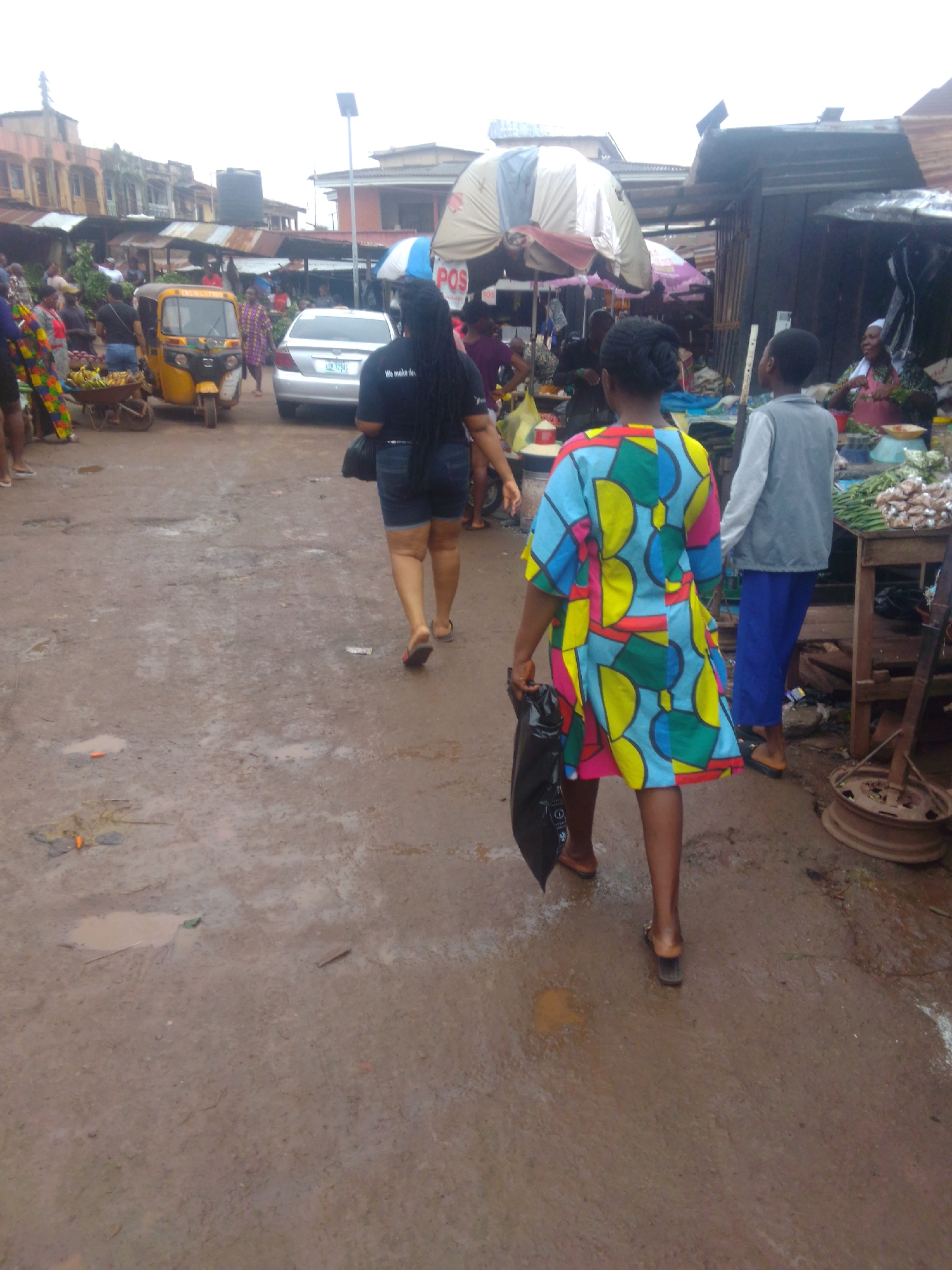 The image depicts a street scene in an outdoor market, possibly in Enugu, Nigeria. The ground is muddy and wet, with puddles scattered across it. A yellow auto-rickshaw is parked on the left side of the frame, with a cart loaded with bananas next to it. In the foreground, two women are walking away from the camera. The woman on the left is wearing a black t-shirt and denim shorts, carrying a black bag. The woman on the right is wearing a brightly colored, patterned dress and carrying a black bag. Behind them, other people are visible, along with market stalls and buildings. The overall impression is that of a busy, somewhat disheveled, and wet marketplace.