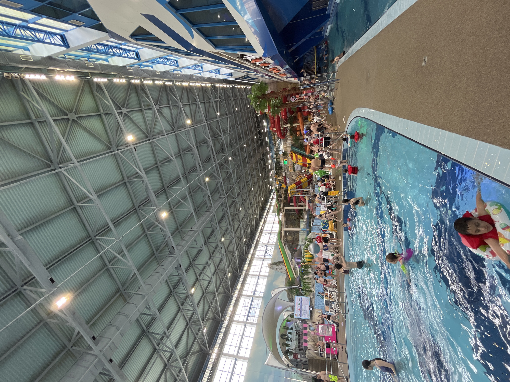The image depicts an indoor water park with a large swimming pool and several water slides. The ceiling is high and features a complex metal structure with lighting fixtures. The walls are decorated with murals depicting natural landscapes, and a large, ship-like structure with blue and white detailing is visible on the right side. Numerous people of various ages are present, some swimming, others relaxing on lounge chairs or standing around the pool area. There are several water slides, including red and yellow ones, and a wave pool area. The overall environment appears to be a recreational facility with a lively atmosphere. This water park is located in the city of Yekaterinburg, Russia.