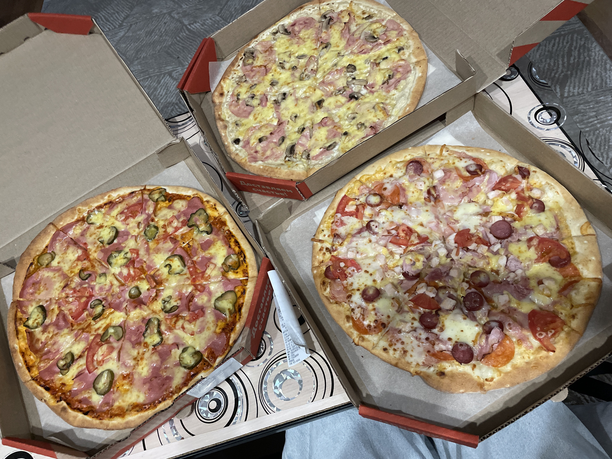 Three pizzas are presented in opened pizza boxes on a grey, patterned surface. The pizza in the top left box is topped with cheese, sliced ham, tomato, and pickles. The pizza in the bottom left box has cheese, sliced sausage, ham, tomato, and onion. The pizza in the right box is topped with cheese, sliced mushrooms, and ham. The scene appears to be indoors, likely in a residential setting. The location is Chaikovsky, Russia.