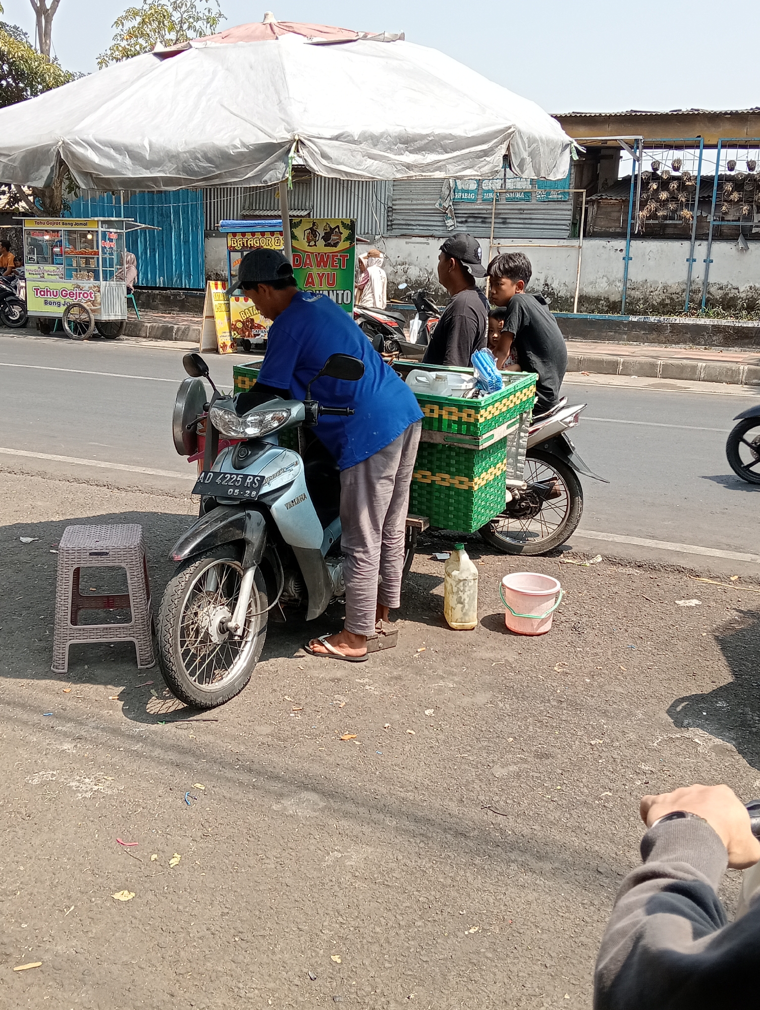 In Grabag, Indonesia, a street vendor stands beside a light blue Yamaha motorcycle parked on the side of a road. The vendor, wearing a blue shirt and gray pants, is attending to the motorcycle. A woven green basket filled with items is attached to the back of the motorcycle. Behind the vendor, two young boys in black shirts are seated on the motorcycle, looking to the side. In the foreground, a small plastic stool and a plastic jug are placed on the ground. A pink bucket is next to the jug. In the background, a large white canopy covers a food stall, and another stall with signs is visible. The road appears to be paved, and the environment suggests a sunny day.
