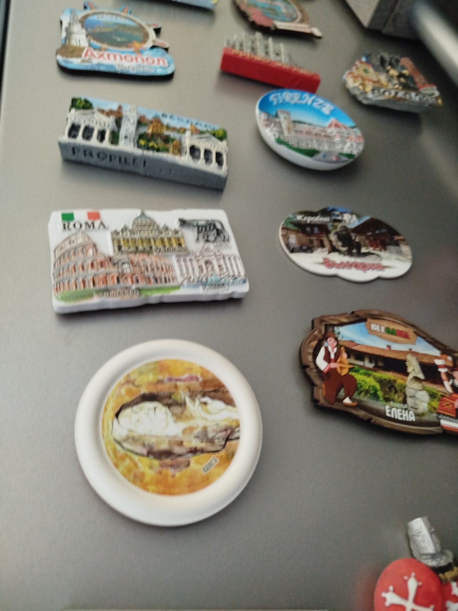 The image shows a refrigerator door covered with various souvenir magnets. There are magnets depicting:
* Axmonon (Likely "Ахмонон" in Cyrillic, a place in Varna, Bulgaria, near Asparuhovo beach)
* Propylaea (Greece)
* Roma (Italy)
* Firenze (Italy)
* Koprivshtitsa, Bulgaria
* Elena, Bulgaria.
There is also a circular magnet with an indiscernible image.
The refrigerator door has a gray surface.