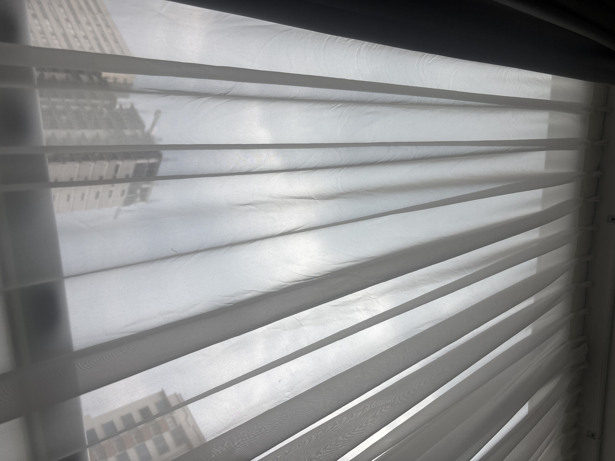The image depicts a view through a window with partially closed white blinds. Through the blinds, a building can be faintly seen. The blinds are angled, creating a series of horizontal lines and shadows. The overall light level appears bright, possibly due to daylight. The location, Jesús del Monte, Mexico, is not discernible from the image alone.