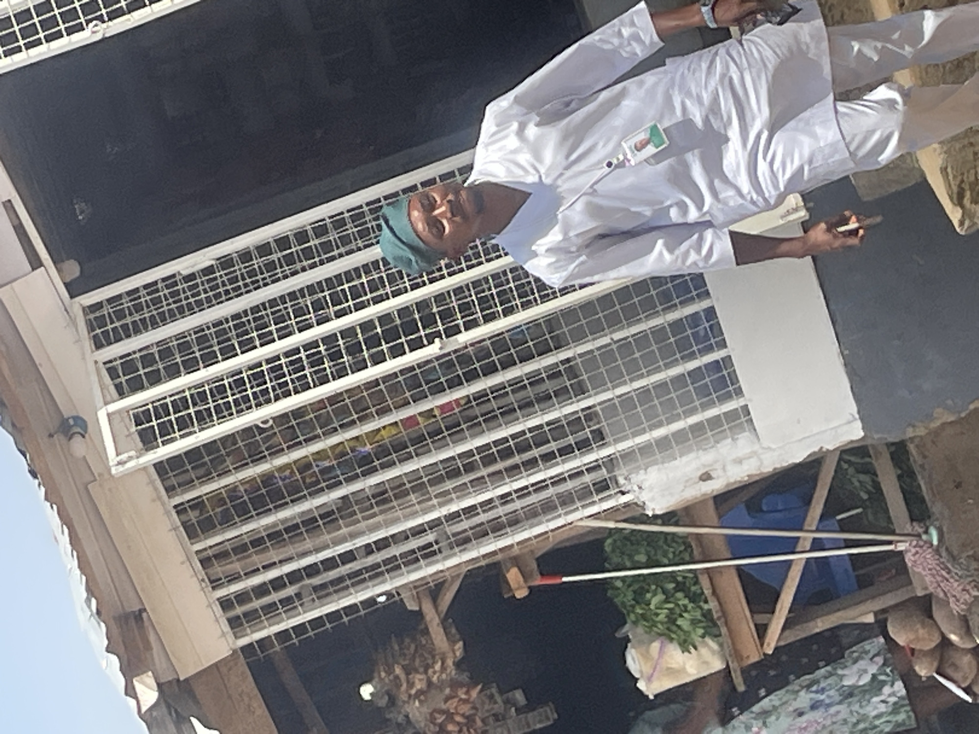 The image depicts a street scene in Abuja, Nigeria, with a man wearing a white traditional outfit and a green cap walking towards the right. He has a lanyard with an identification card around his neck, which features a picture of him. In the background, there is a stall with metal bars and a corrugated iron roof, where various items are displayed, including produce and other goods. A mop with a pink handle lies on the ground in front of the stall. The scene suggests a busy marketplace or commercial area typical of an urban environment in Nigeria.