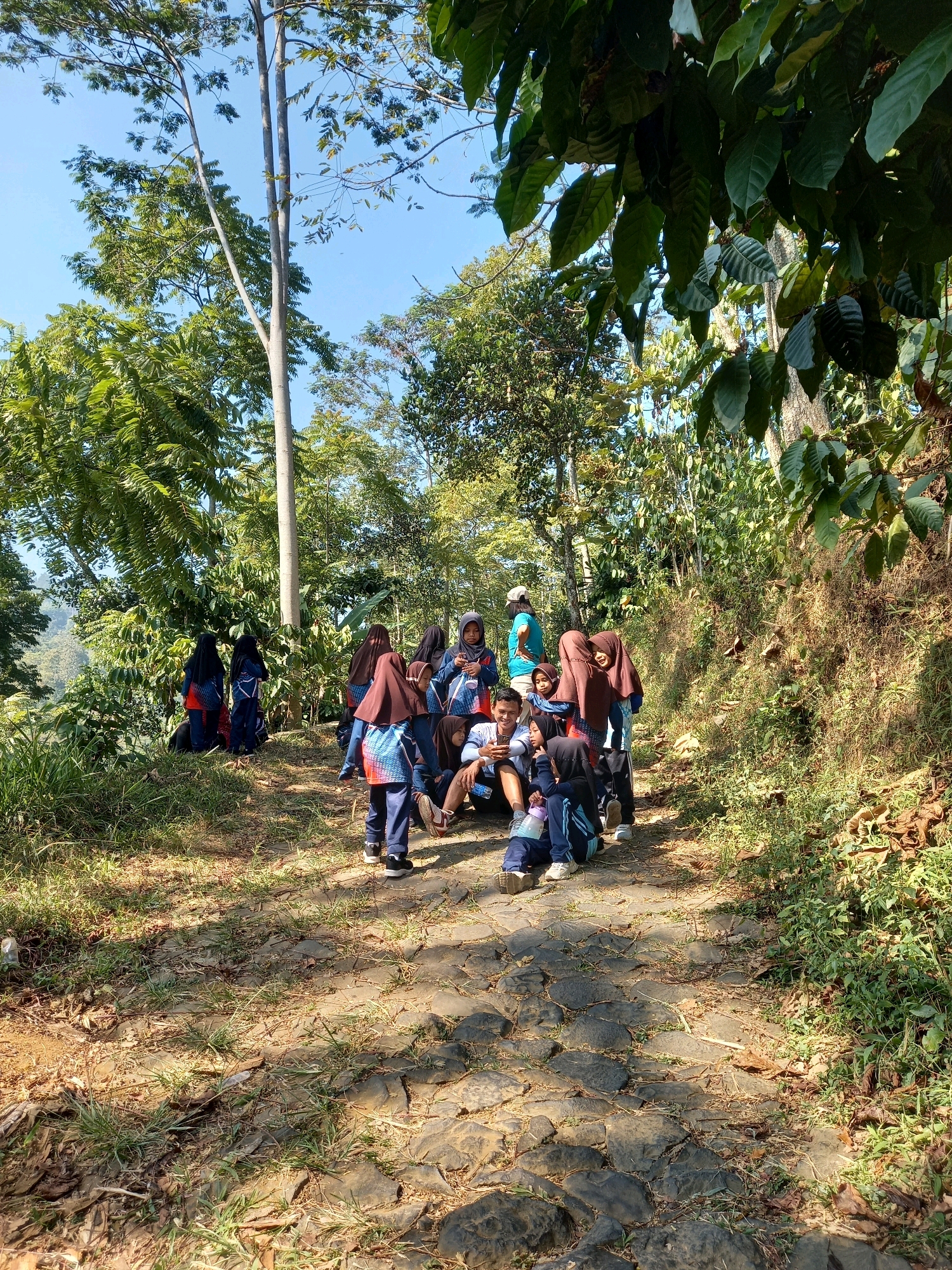 A group of approximately ten children and young adults, some wearing hijabs, are gathered on a cobblestone path in a rural, forested area. The group appears to be taking a break, with some sitting on the ground and others standing and interacting. One individual in the center is looking at a mobile phone. The environment is characterized by lush green vegetation, tall trees, and a clear blue sky. The path is uneven and bordered by dry leaves and grass. The overall impression is a casual gathering in a natural, outdoor setting. The location is identified as Blumah, Indonesia.