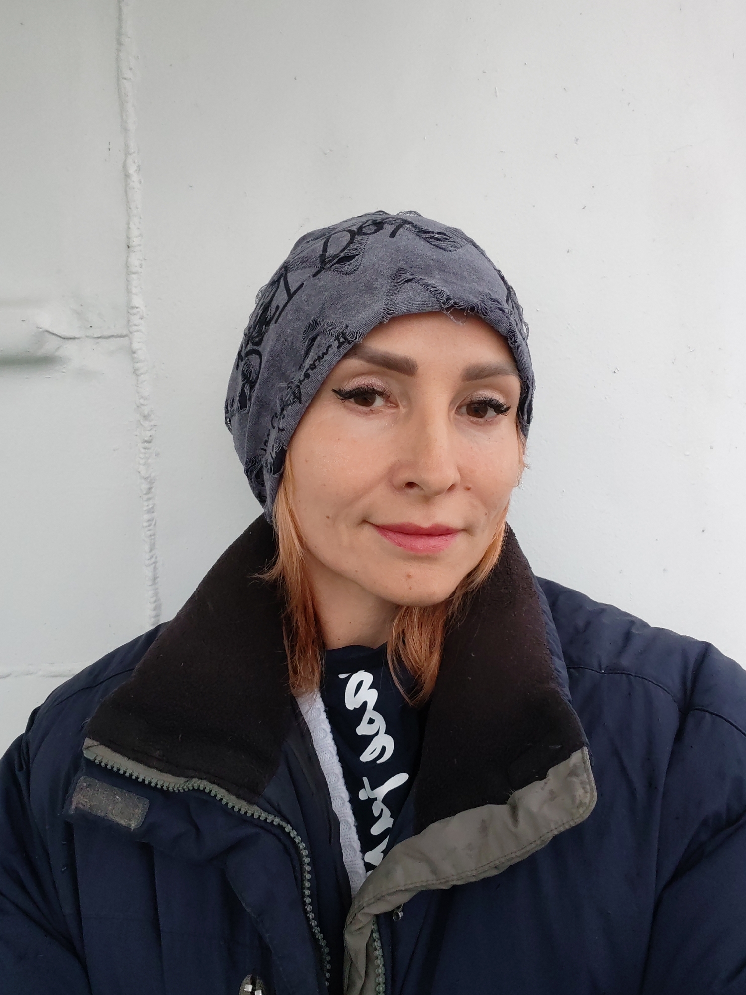 A woman wearing a blue jacket and a grey beanie stands against a white wall. The wall has a vertical seam running up the center. She has blonde hair and is wearing makeup, including eyeliner and lipstick. A dark t-shirt with white lettering is visible beneath her jacket. The location is stated to be Goritsy, Russia.