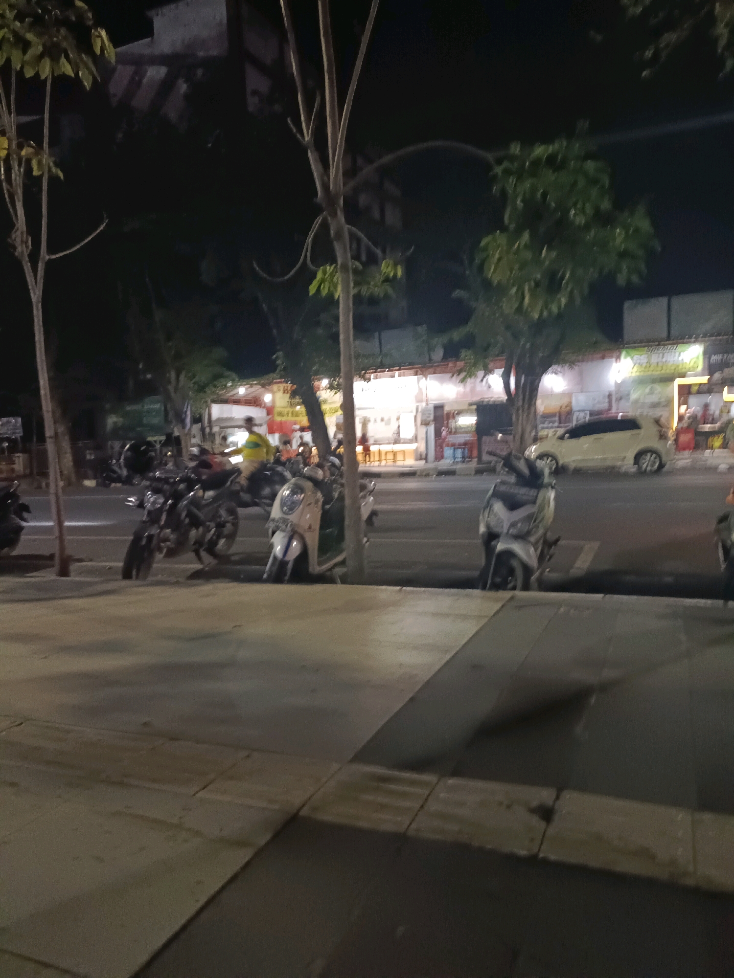 The image depicts a nighttime street scene in Lamongan, Indonesia, likely near a row of shops. A sidewalk with large concrete tiles is in the foreground, casting shadows from streetlights or nearby lamps. Across the street, several parked motorcycles are visible, some with riders. A white car is also on the road. Behind the motorcycles and car, a commercial strip with illuminated storefronts can be seen, some of which appear to be food vendors or small businesses. Trees line the street, adding to the urban landscape. The overall atmosphere suggests a busy evening, common in Indonesian cities.