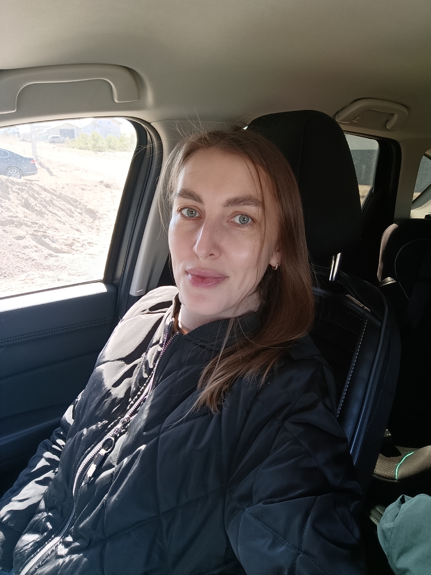 A woman is sitting in the driver's seat of a car. She has light skin, light brown hair, and blue eyes. She is wearing a black quilted jacket over a beige turtleneck. The car is parked on a dirt road near a construction site. In the background, there are houses and other cars. The location is near Chaikovsky, Russia.