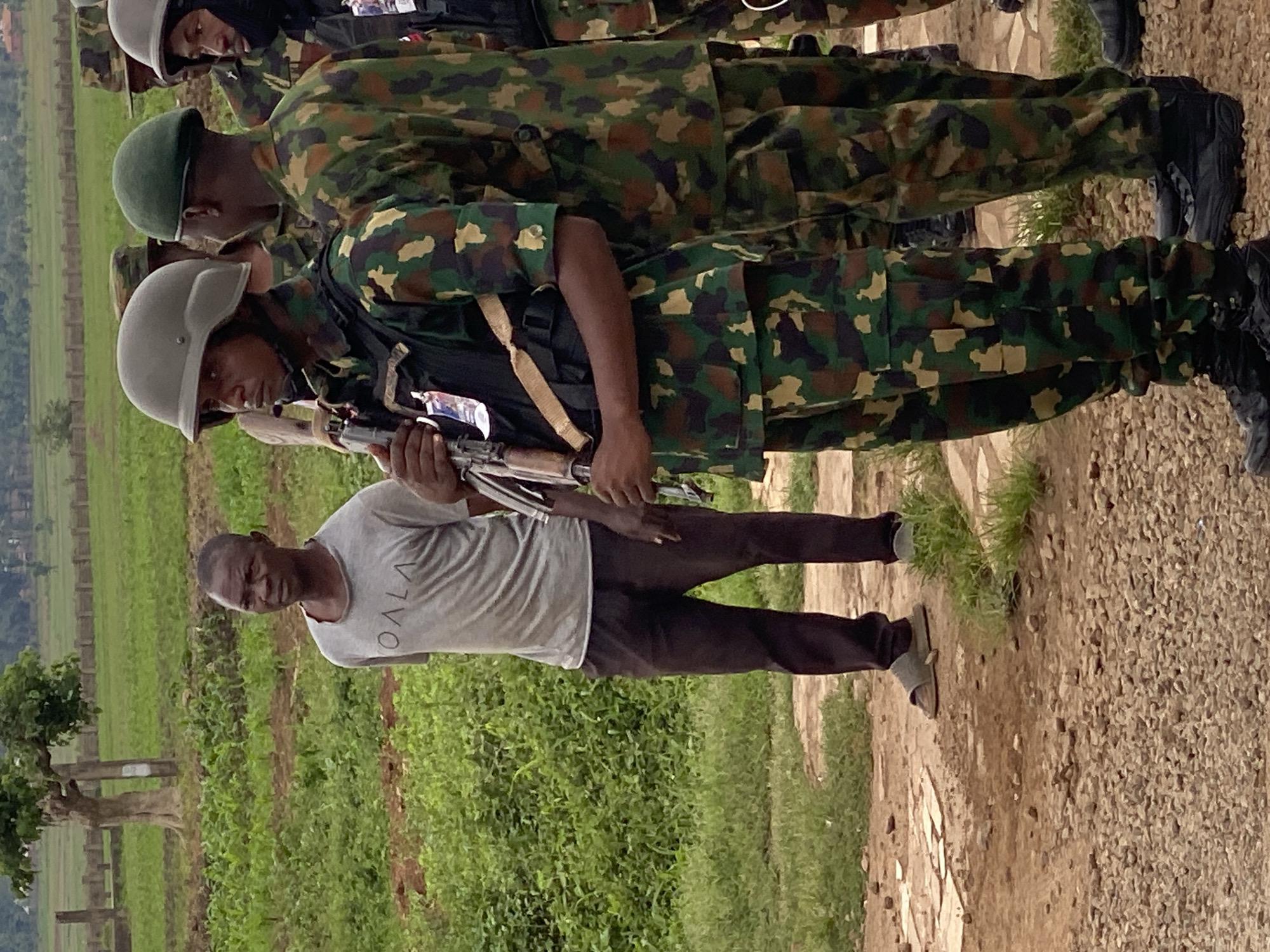 A low-angle, outdoor shot shows a single African male, wearing a grey t-shirt and dark trousers, holding an AK-47 rifle. He is positioned in front of two soldiers wearing camouflage fatigues and helmets. The area is a gravel patch next to a grassy field. A row of trees and a fenced area are visible in the background. The location is in the city of Maigizo, Nigeria.