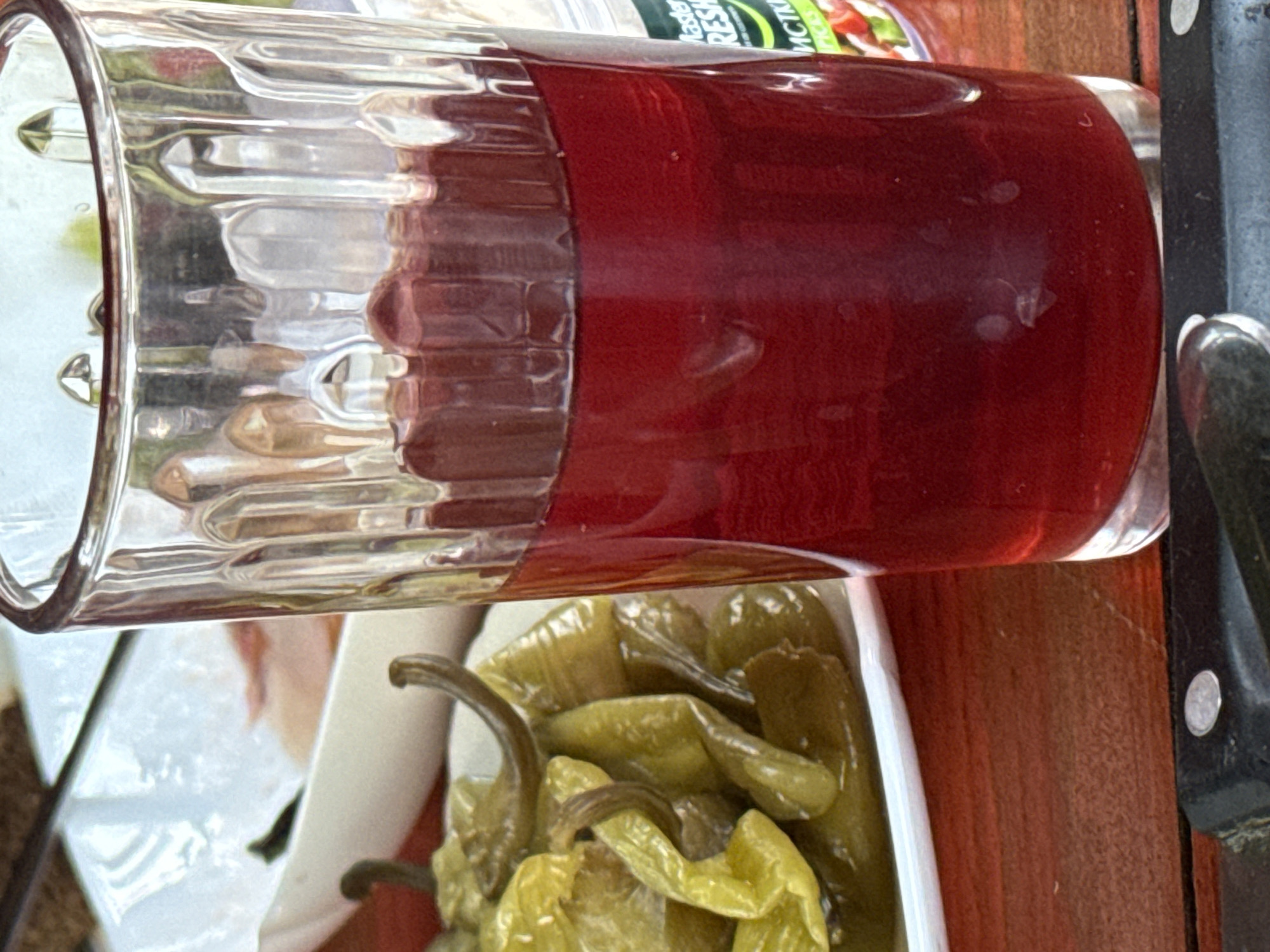 A close-up shot shows a glass filled with a dark red beverage, possibly juice or wine, placed on a wooden surface. Next to the glass, on a white plate, are pickled green peppers. In the background, there's a hint of other food items and what appears to be a jar of pickles with a label. The scene suggests a meal setting, possibly outdoors due to the lighting and the wooden table. The image is taken in Kerch, Ukraine.