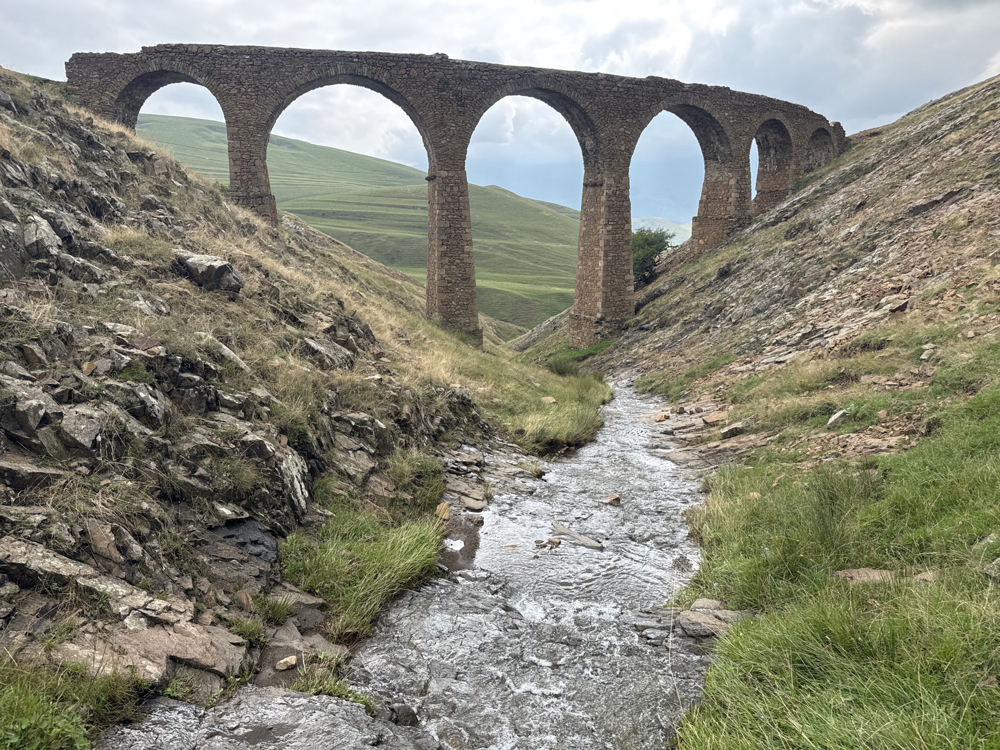 The image captures an ancient stone aqueduct with multiple arches spanning a narrow, rocky ravine. A shallow, fast-flowing stream runs through the bottom of the ravine, with grass and rocks lining its banks. The surrounding landscape is hilly and green, with a cloudy sky overhead. The aqueduct is made of rough-hewn stones, showing signs of age and weathering. The terrain is rugged and natural, suggesting a remote or undeveloped location. The scene appears to be located in Miskinli, Azerbaijan, a region known for its historical ruins and natural beauty.