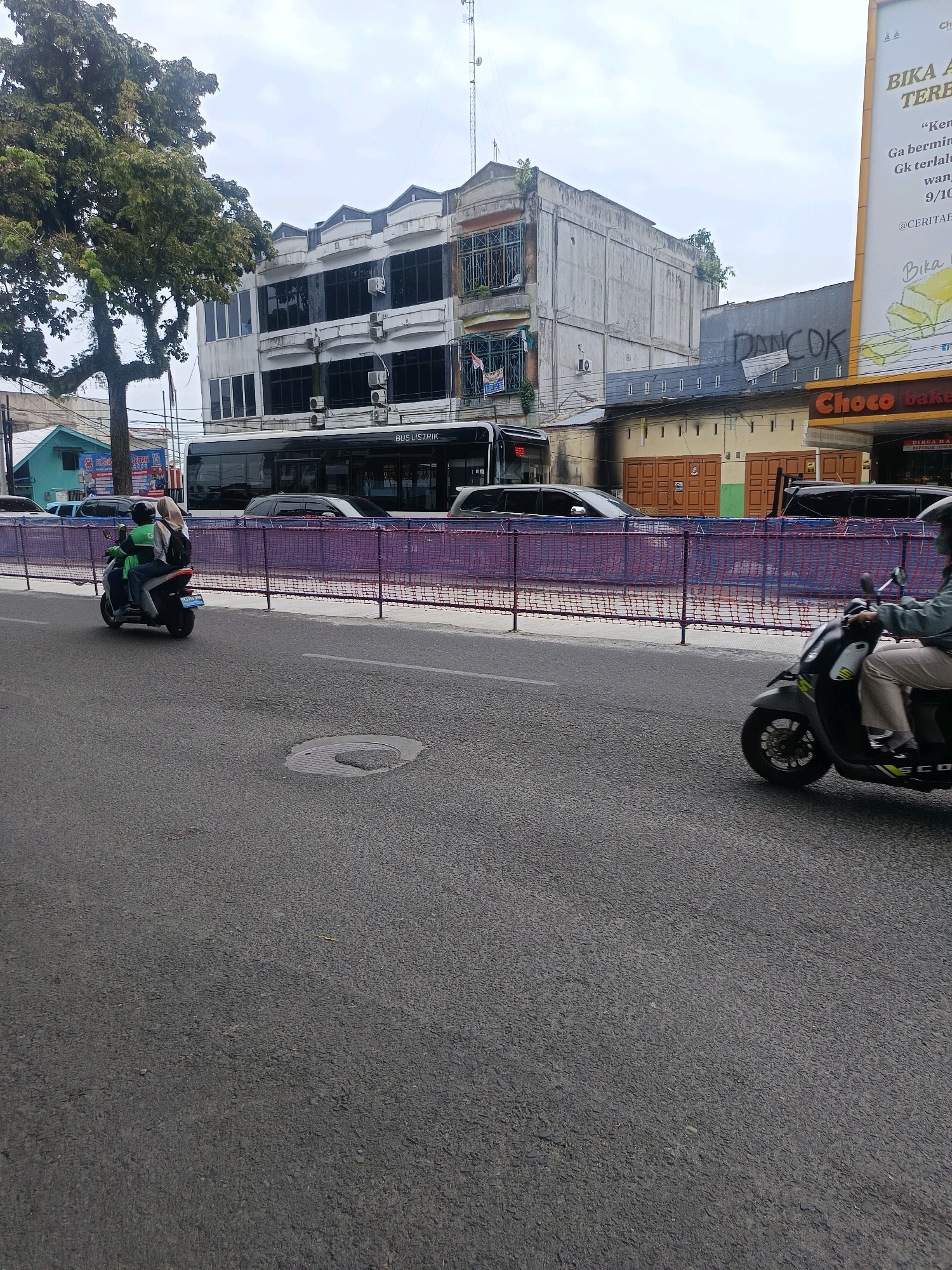 In the city of Medan, Indonesia, a two-wheeled motor vehicle travels on an asphalt road. A male rider, wearing a blue jacket and a dark helmet, is accompanied by a female passenger who wears a beige headscarf and a backpack. Both individuals are seated on the motor vehicle. The motor vehicle is traveling on the right side of the road, and there is a purple safety fence installed on the left side of the road. In the background, there is a double-decker bus, and several other vehicles. Above the road, there are several buildings, some of which are multi-story. The sky is overcast with clouds. The buildings are of various architectural styles, and some appear to be commercial establishments. The overall scene depicts a typical urban streetscape in Medan.