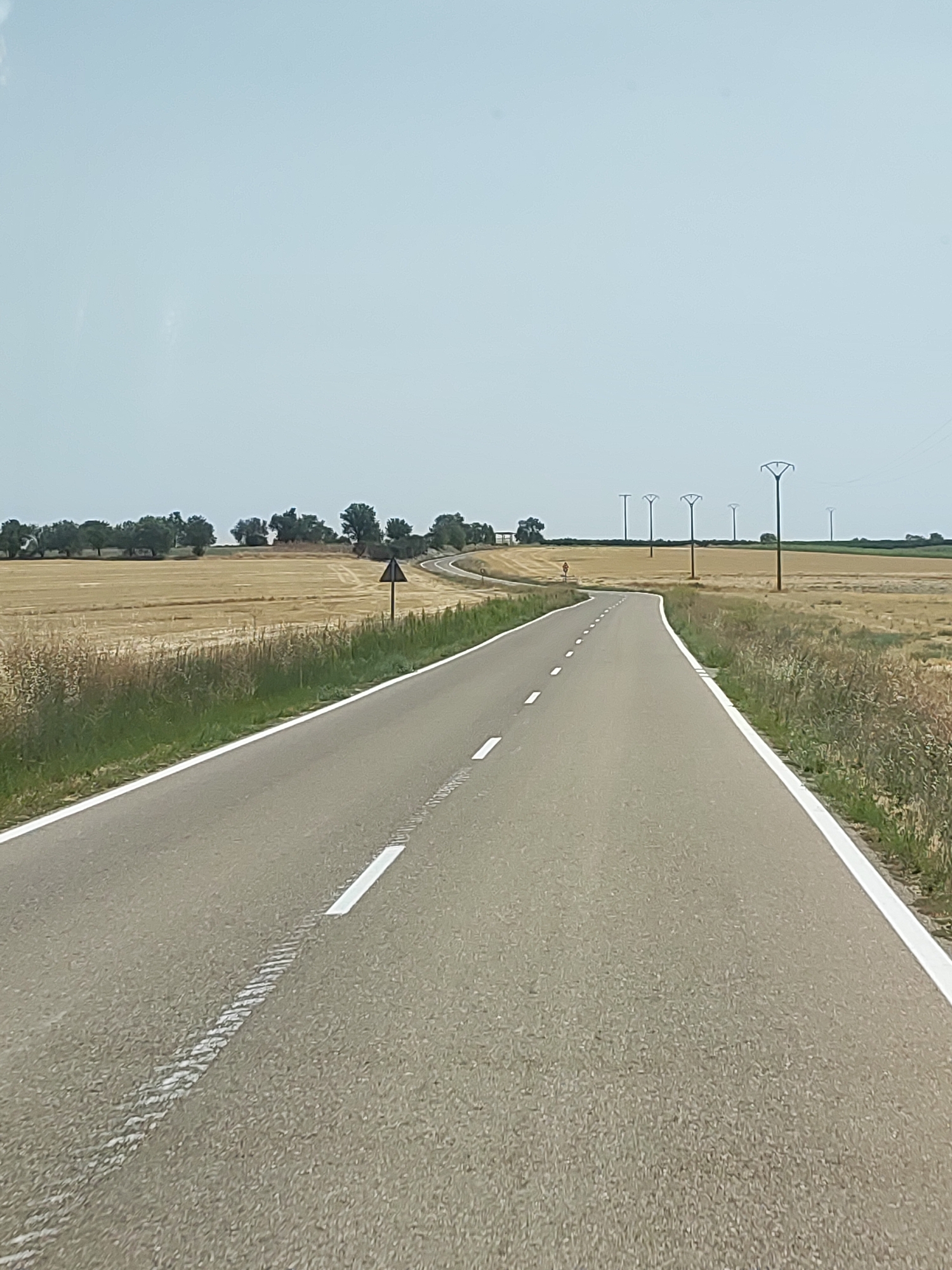 The image depicts a two-lane asphalt road winding through a rural landscape. The road is marked with white lines, a solid line on the right edge and dashed lines dividing the lanes. To the left of the road, a field of harvested grain, likely wheat or barley, extends to a line of trees in the distance. To the right, a similar field of harvested grain is visible, bordered by dry grass and vegetation. Several tall electricity pylons are sparsely distributed across the fields. A triangular warning sign is positioned on the left side of the road, near a curve. The sky is overcast with a pale blue hue. The scene appears to be in the municipality of Cigales, Spain, given the characteristic flat, agricultural landscape often found in the region, and the style of the road and utility infrastructure. There are no people or vehicles visible in the image.