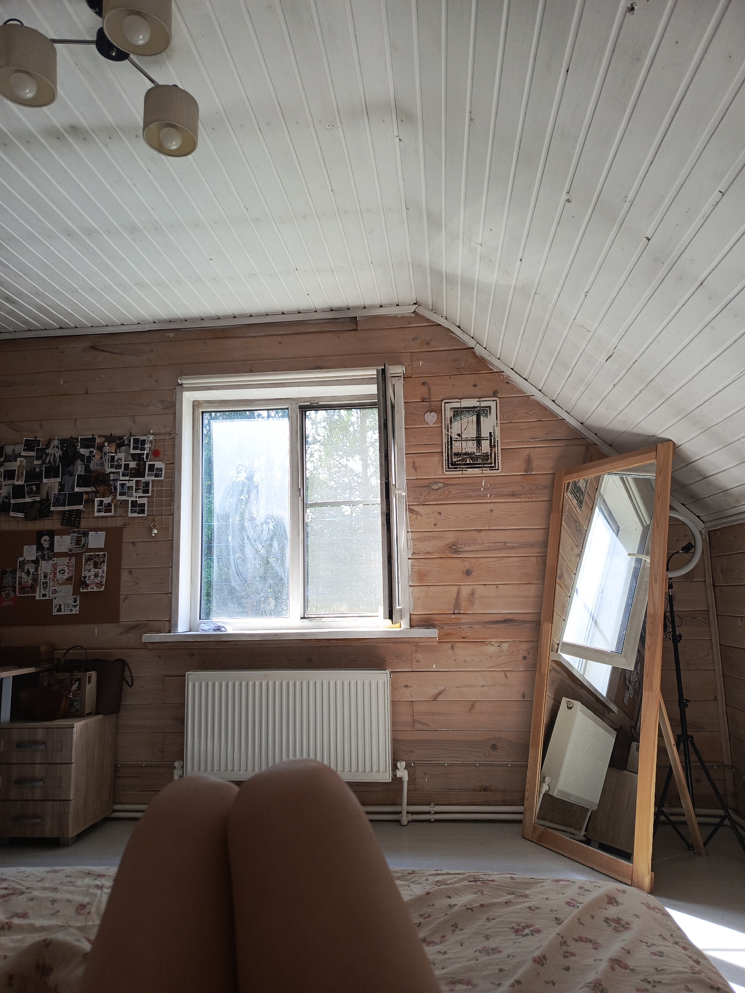 A room in Северный, Russia, has wood paneling on the walls and ceiling. A light fixture with three shades is visible in the upper left. A window with a white frame is slightly ajar. A radiator sits beneath the window. To the left of the window is a bulletin board with photographs and papers. To the right, a full-length mirror leans against the wall, reflecting the radiator. A ring light on a tripod stands behind the mirror. A bed with a floral patterned cover is visible in the foreground with a pair of legs at the very front.