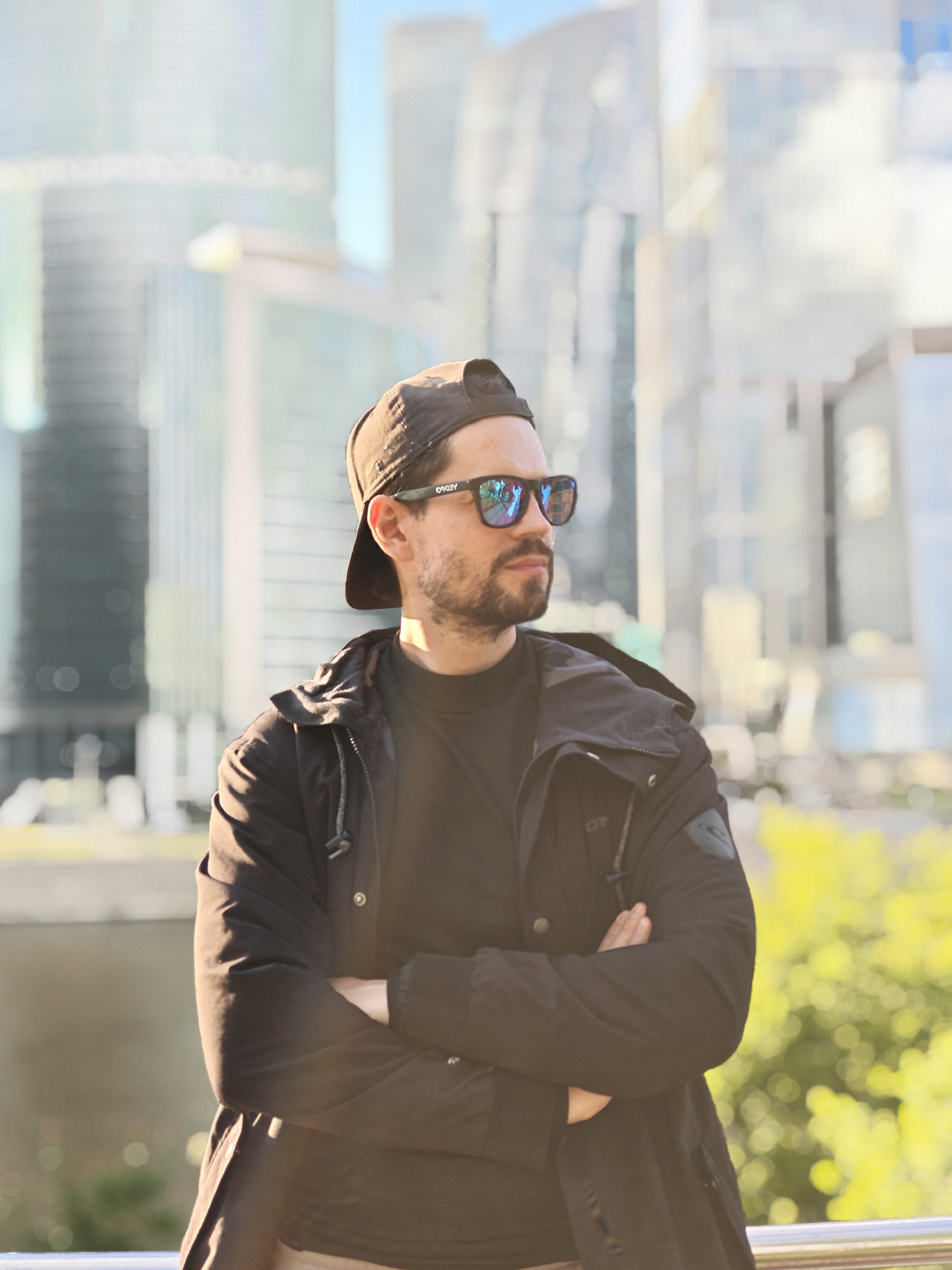 A medium shot depicts a bearded man with reflective sunglasses and a black baseball cap facing right with crossed arms. He is wearing a black jacket over a black shirt. The background features modern glass skyscrapers and green foliage, indicative of an urban environment. The location appears to be in Moscow, Russia, suggested by the architectural style of the buildings in the background, which resemble those in the Moscow International Business Center (Moscow City).