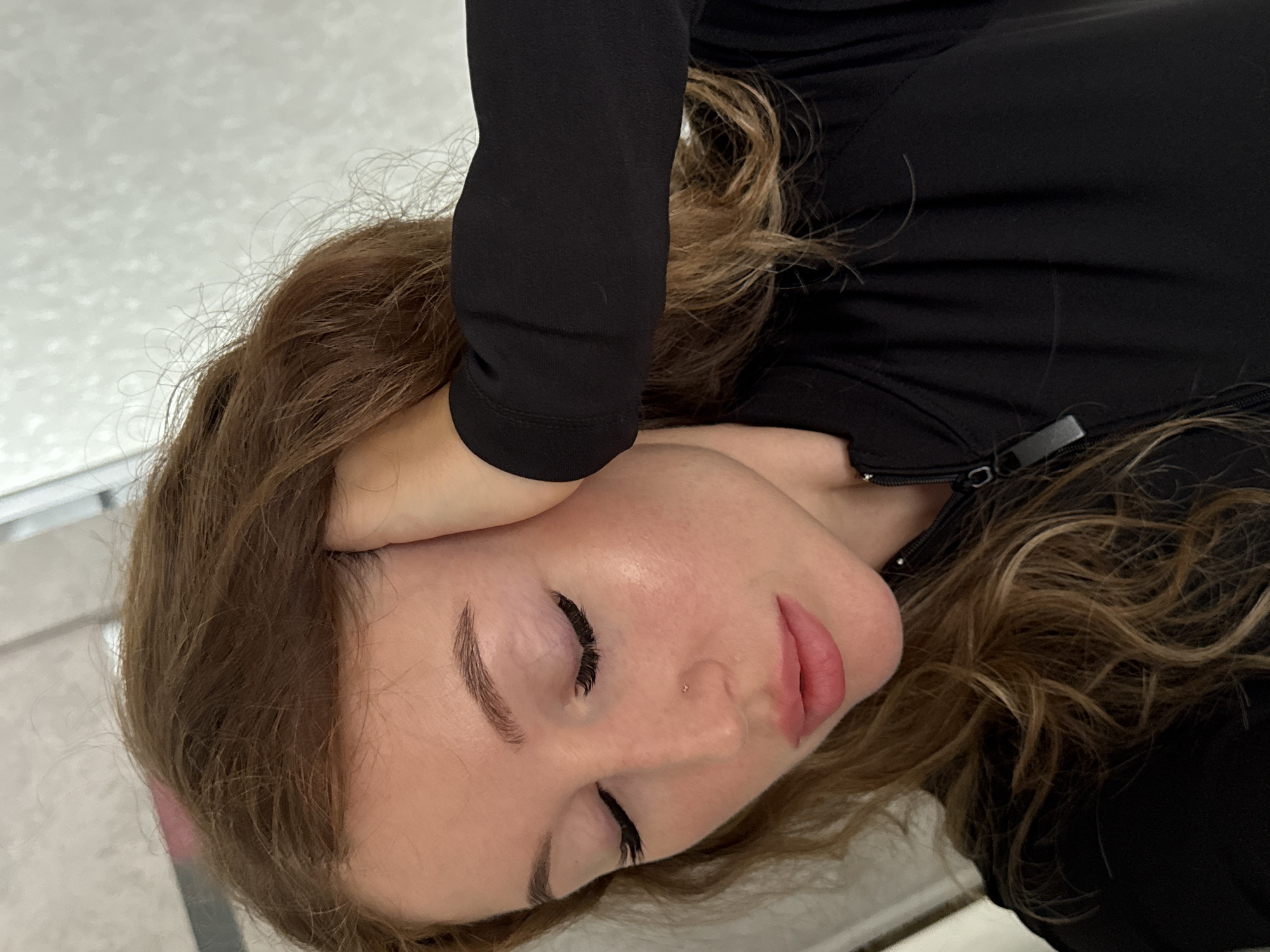 A woman with light skin and light brown hair, wearing a black long-sleeved garment, lies on a surface of an indeterminate material. Her eyes are closed. A hand, covered in black fabric, rests on her forehead. The setting appears to be indoors, with a neutral-colored background. The image provides no indication of being in the city of Chaikovsky, Russia.