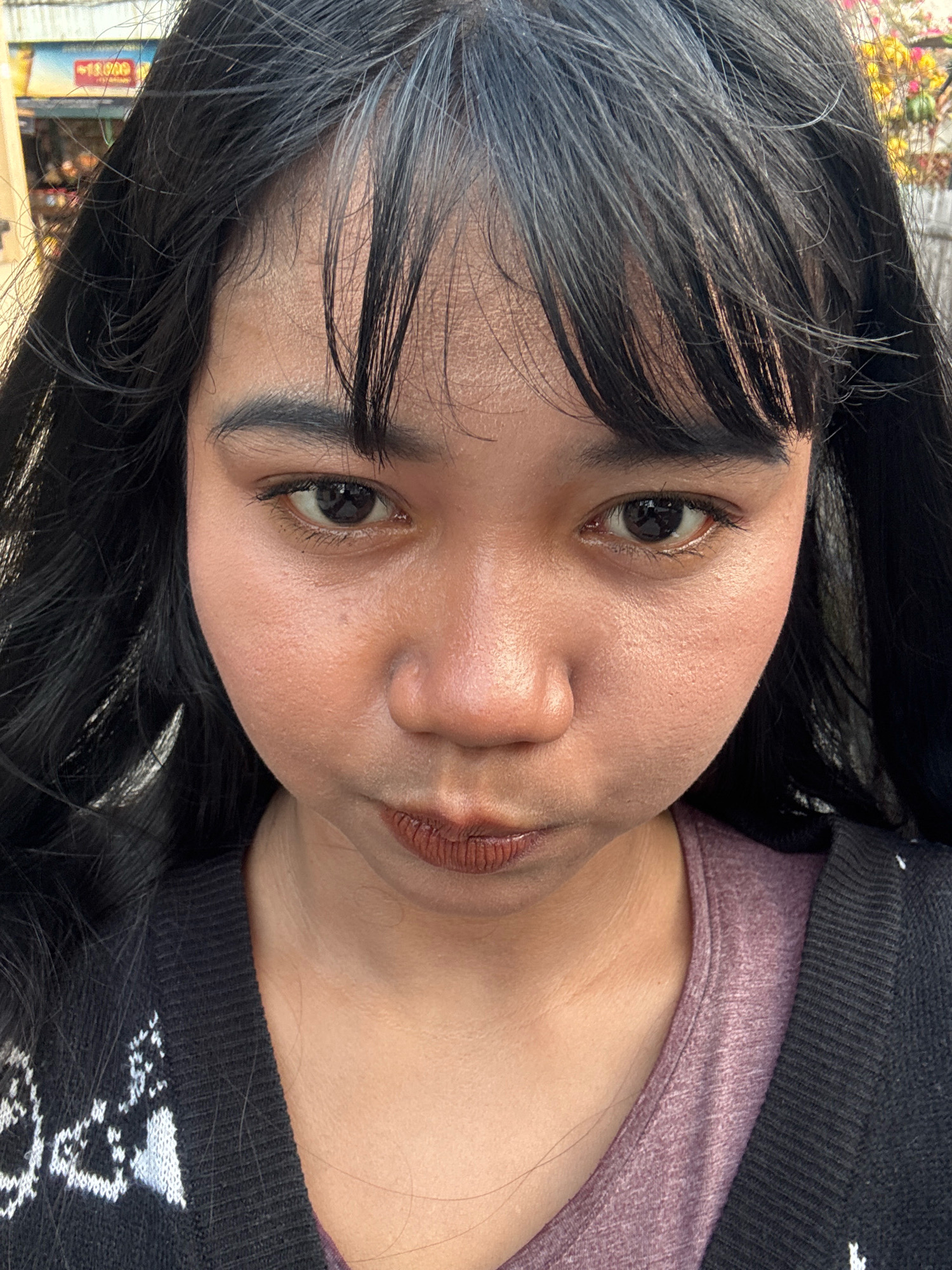 A medium shot, frontal, eye-level photograph shows a young Indonesian woman with long, dark hair, some strands of which are graying. She has dark eyes, thin eyebrows, and is wearing dark lipstick. Her skin is a warm, medium tone, and she appears to be wearing a dark cardigan over a purple shirt. The background is slightly blurred, showing a building with colorful signage and some foliage. The setting is the street in North Jakarta, Indonesia, specifically in front of a shop in the Ancol area. The overall impression is of a casual, outdoor portrait.