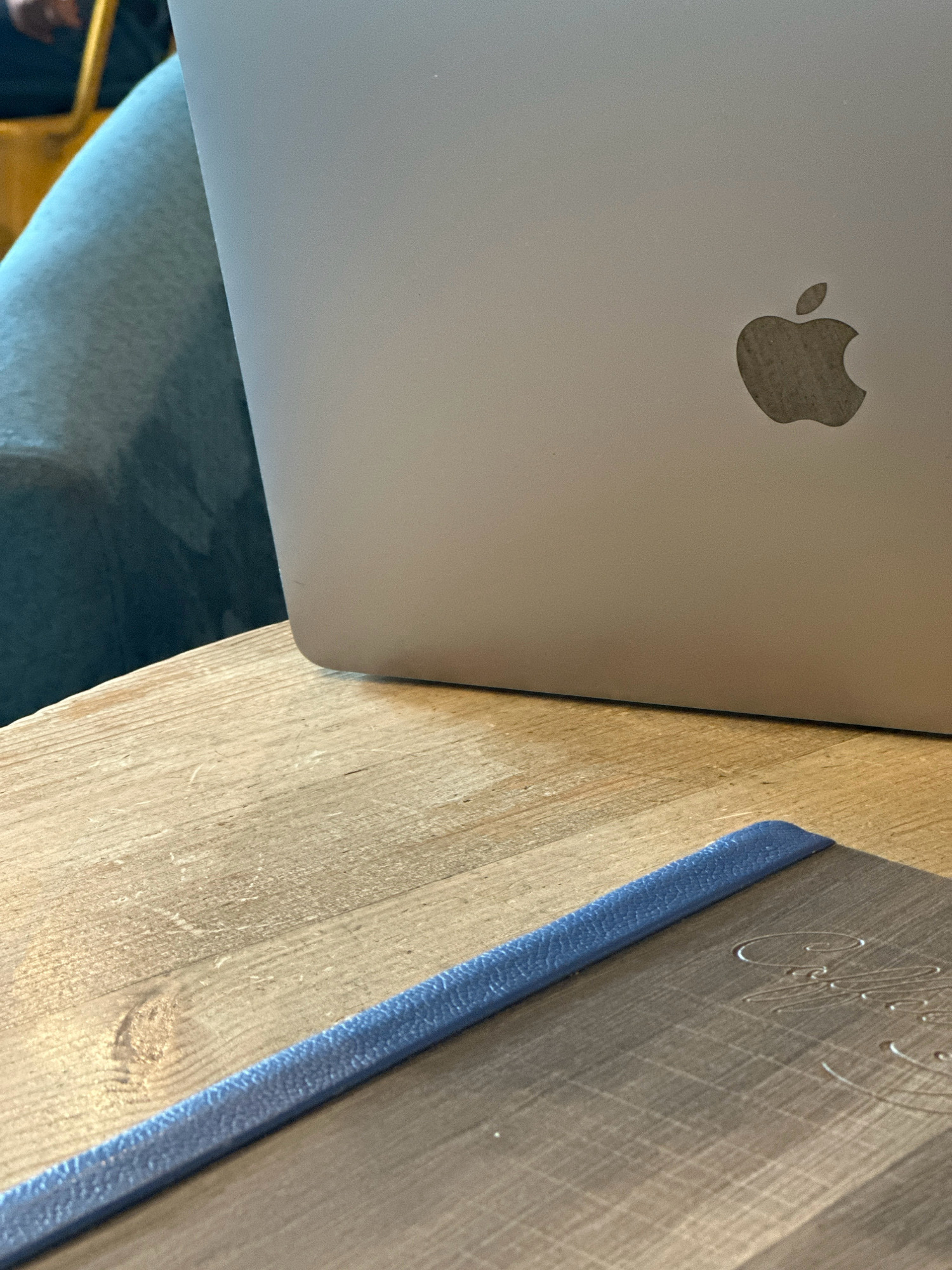 A close-up, eye-level shot shows the Apple logo on a silver MacBook laptop. The laptop is positioned on a wooden table next to a blue-covered notebook. In the background, a person's legs are visible, clad in light blue pants, seated on a yellow chair. The lighting suggests an indoor setting, possibly a cafe or study area. The image does not provide any specific geographical indicators to determine its location within Kathmandu Metropolitan City, Nepal.