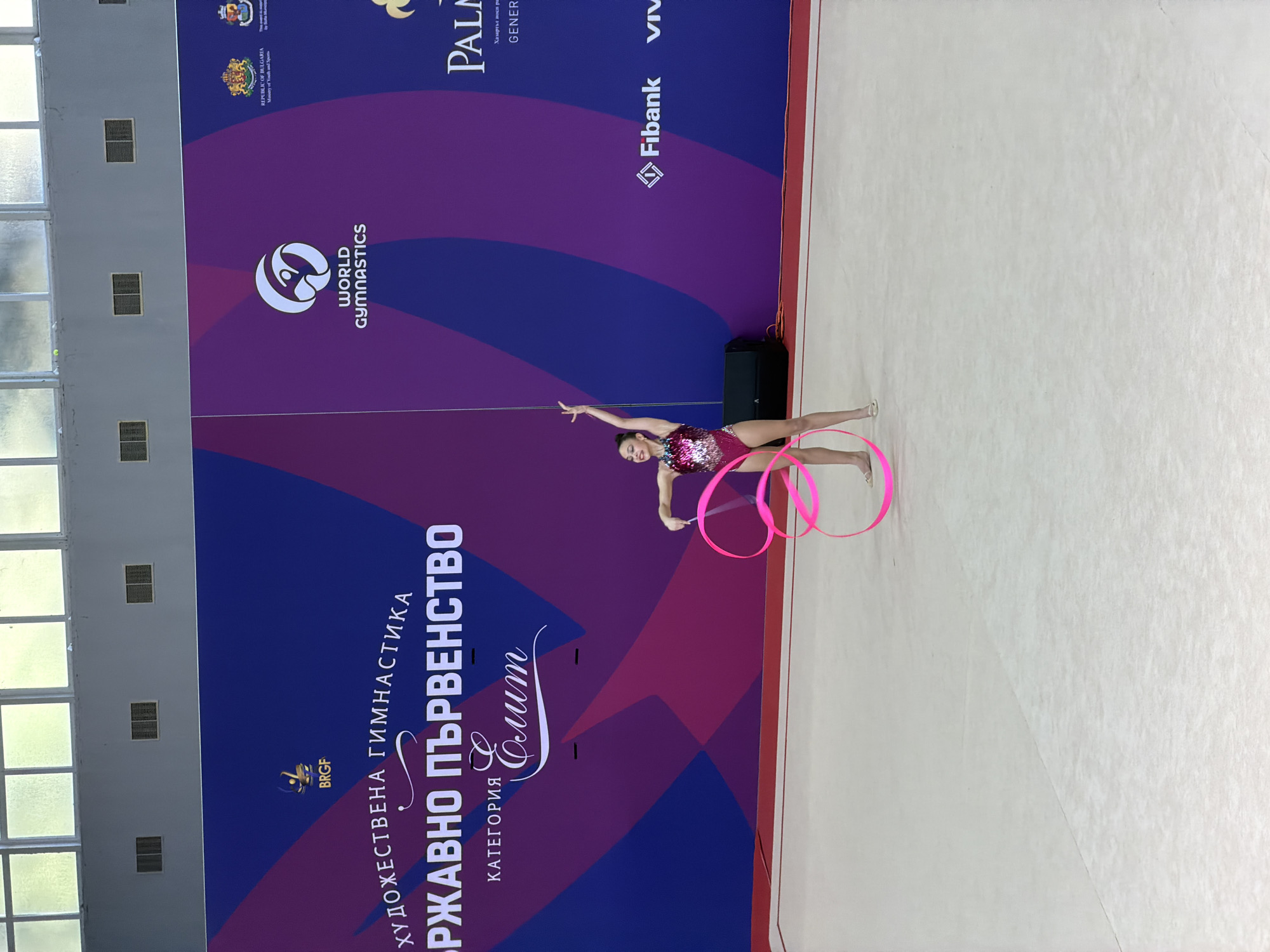 Here is a description of the image:
The image shows a rhythmic gymnast performing with a ribbon and hoops at a competition. The gymnast is wearing a sequined leotard and is positioned on a white floor. In the background is a banner with the logos of the competition and sponsors. The banner includes text in Bulgarian, identifying the competition as the "Държавно първенство" (State Championship) in rhythmic gymnastics, category "Елит" (Elite). The World Gymnastics logo is also visible. Sponsors include Fibank and Viva. The competition appears to be held in Sofia, Bulgaria, as evidenced by the text "REPUBLIC OF BULGARIA Ministry of Youth and Spans" on the banner.