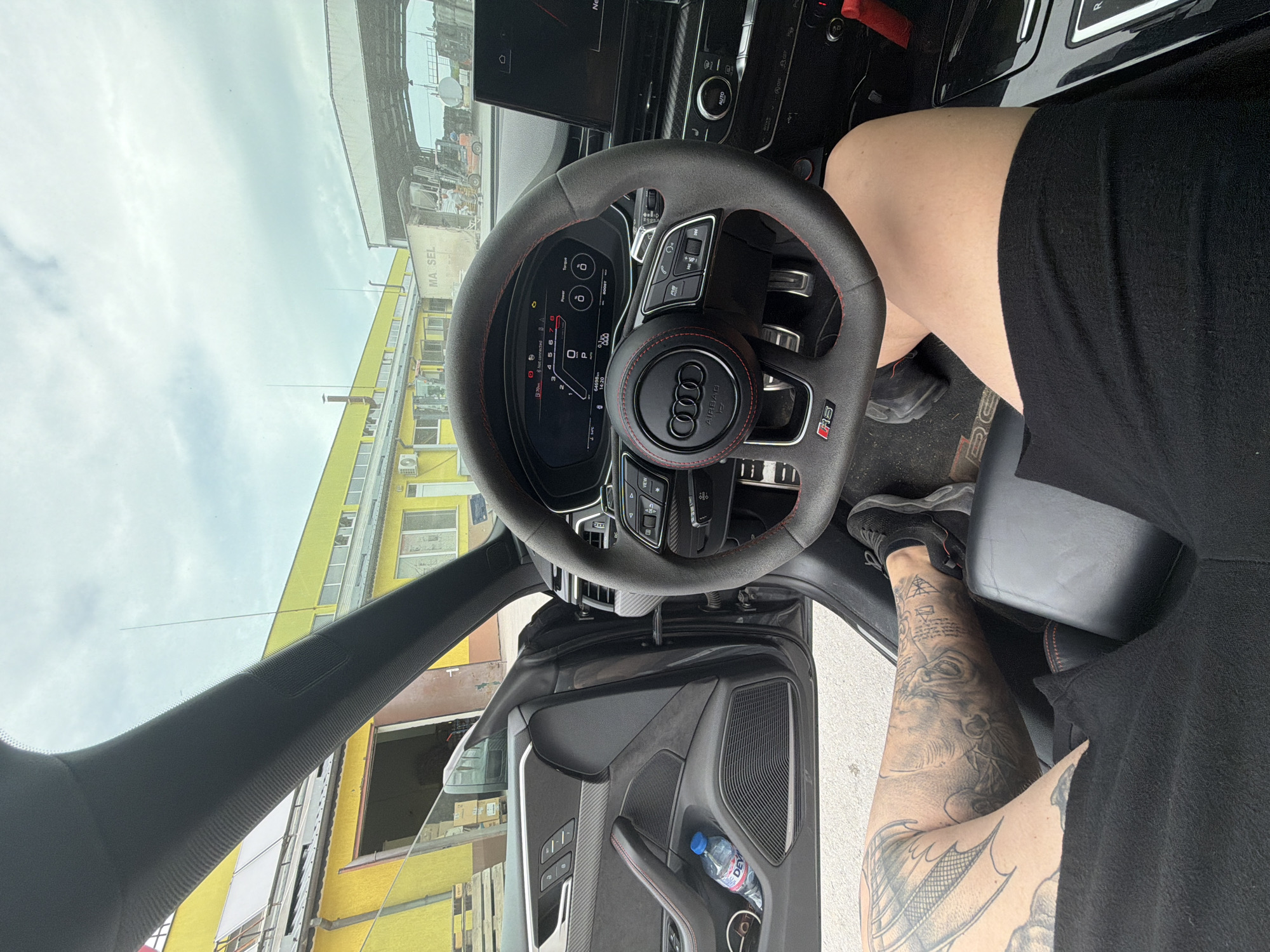 A first-person perspective image shows the interior of a black Audi RS vehicle. The steering wheel prominently features the Audi logo and red stitching. A person wearing black shorts and shoes is in the driver's seat, with a visible tattoo on their lower leg. A water bottle is present in the car door. The exterior view through the windshield and open door shows a yellow building, suggesting a location in Dolno Ablanovo, Bulgaria, based on the MARSEL label visible on the building. The scene appears to be taken during daylight hours.