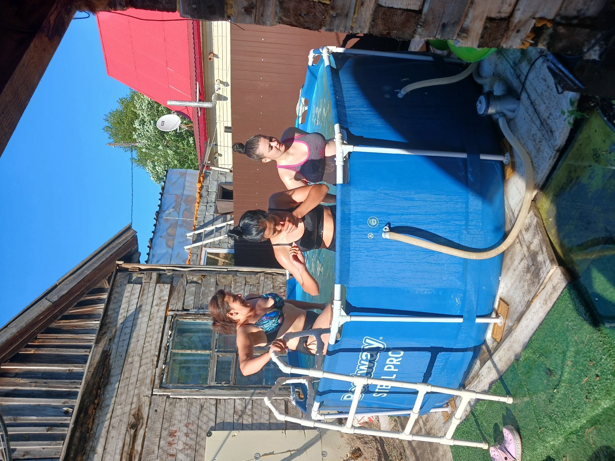 In Chik, Russia, on a sunny day, three young women are in a blue, above-ground swimming pool. The pool is labeled "Bestway Steel Pro." One woman, with brown hair in a ponytail, wears a floral bikini and is standing, reaching for the pool's edge. Another woman, with dark hair pulled into a bun, wears a black sports bra and black bottoms, her face partially obscured as she appears to be splashing water. The third woman, with her hair in a bun, wears a grey and pink sports bra and dark bottoms, looking down into the water. The pool is set up on a concrete base with wooden supports. A white ladder provides access to the pool. In the background, a rustic wooden building stands to the left, with a weathered facade and a window. To the right, a brown wooden fence separates the property from other structures, including a building with a bright red roof and a satellite dish. Green grass and some overgrown vegetation are visible around the pool area. A small water pump with hoses is connected to the pool.