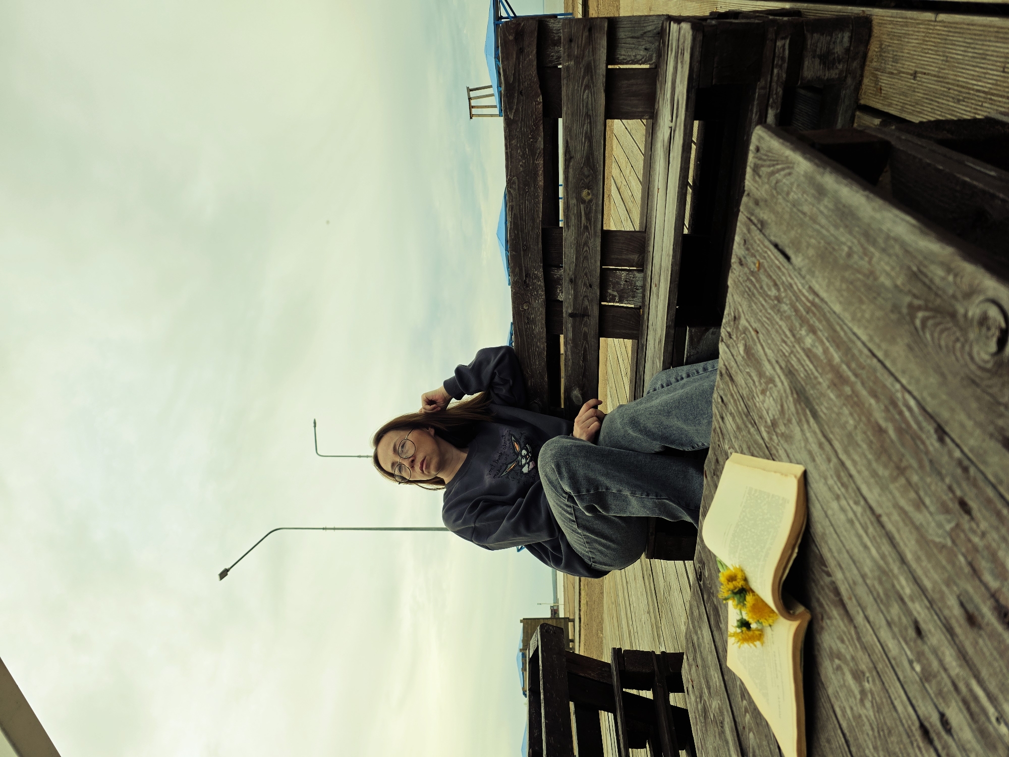A woman with glasses is sitting on a wooden bench, with her hand in her hair. She is wearing a dark-colored sweatshirt and blue jeans. On the table to her right, an open book with yellow flowers rests. In the background, there is a beach and a cloudy sky. There are street lights and wooden structures in the distance. The image appears to have been taken in Chaikovsky, Russia.