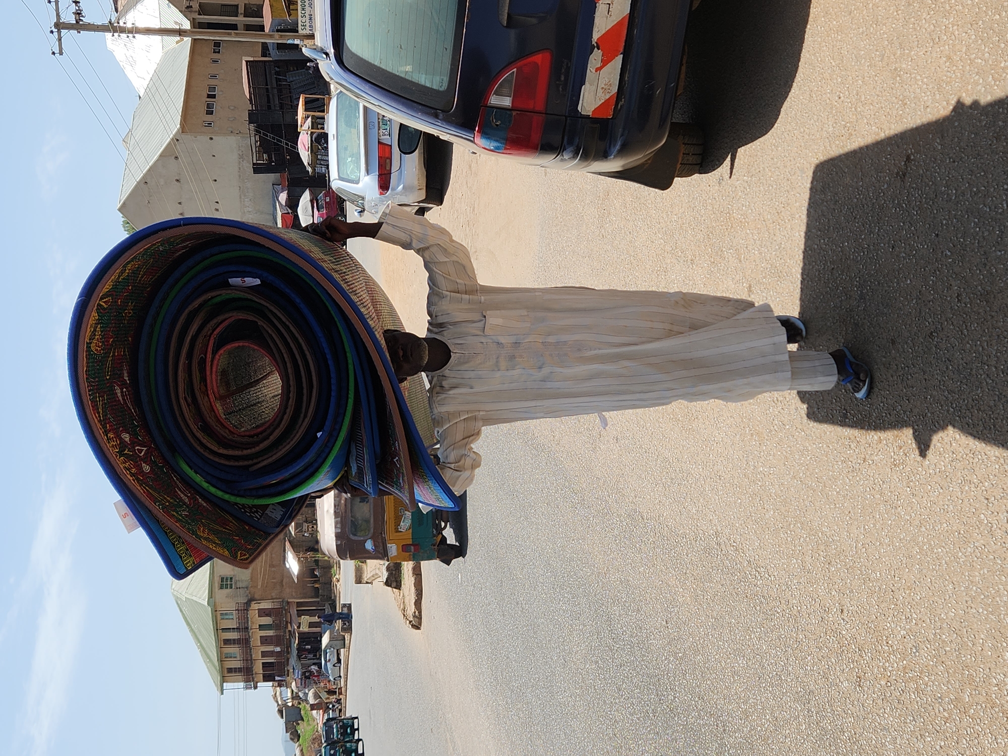 A street scene in the city of Unknown, Unknown features a man carrying a stack of rolled mats on his head, walking on a paved road. The mats are varied in color and texture, with visible rolled edges in blue, green, and brown. The man is wearing a light-colored, striped robe and blue sneakers. A parked car is partially visible in the upper right corner of the frame. Buildings are seen in the background, along with other vehicles and pedestrians at a distance. The scene is brightly lit, with a clear sky.