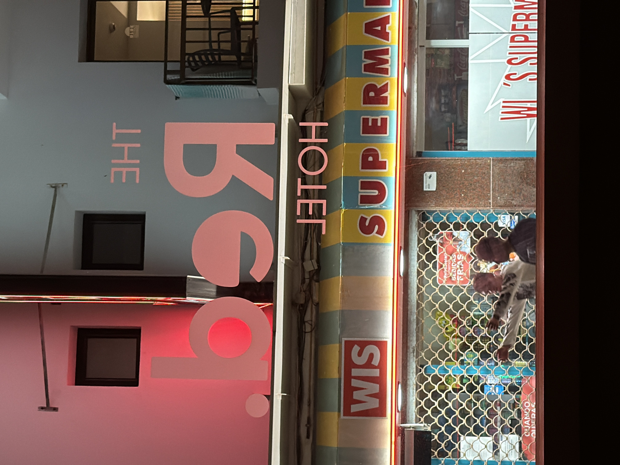 This is a vertical, slightly tilted photograph of a storefront in Sant Antoni De Portmany, Spain. The storefront features a colorful, striped facade with large, pink lettering that reads "THE BG". To the right of "THE BG", the word "HOTEG" is visible in pink letters, followed by "SUPERMA" in red and yellow lettering. The store appears to be a supermarket, as indicated by the text "WI 'S SUPERM" on a white sign to the right of the entrance.
Through a metal grate, two individuals are visible inside the store. One person, wearing a white sweatshirt with a graphic print, is reaching towards a shelf. The other person is partially obscured. Shelves stocked with various products are visible behind them. Red neon lights line the edge of the storefront. Some signs within the store are in Spanish, including "BERIDAS FRIAS" (cold drinks) and "CUANDO QUIERAS" (whenever you want). The overall impression is a brightly lit, possibly bustling, local shop. The time of day is unclear, but the lighting suggests it could be evening or nighttime.