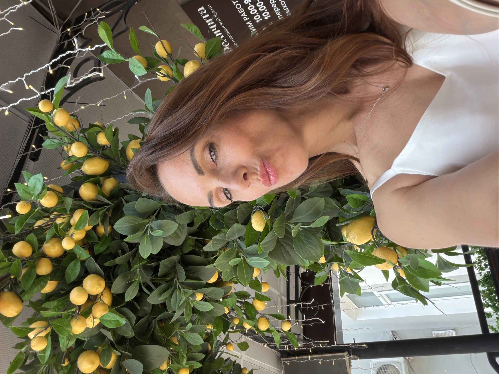 A woman with auburn hair and a white top is taking a selfie. She is pouting her lips and looking at the camera. Behind her, there is a decorative lemon tree with many yellow lemons and green leaves. String lights are visible in the background, as well as a building with windows and an air conditioning unit. A sign in Russian is also visible on the building. The setting appears to be outdoors, possibly a cafe or restaurant patio. The specific location in Birobidzhan, Russia is not discernible from the image.
