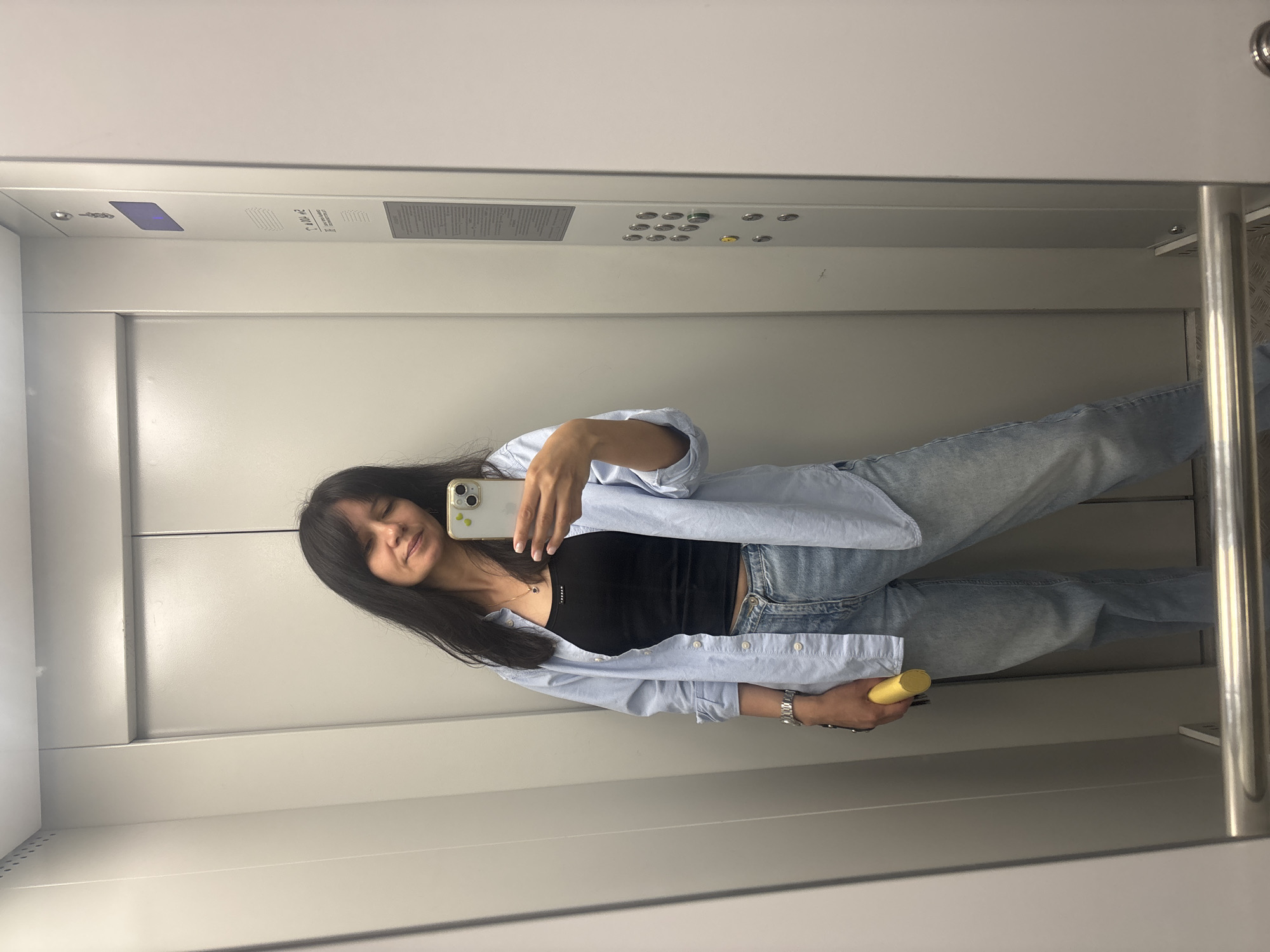 A young woman is captured taking a selfie in a reflective surface, likely an elevator mirror. She has long dark hair with bangs, fair skin, and a relaxed, pleasant expression with a subtle smile and eyes gently closed. She is dressed in a light blue button-down shirt worn open over a black tank top and light blue jeans. A silver watch is visible on her left wrist, and she wears a delicate necklace with a small pendant.
She holds a light-colored smartphone, possibly an iPhone with a case featuring small green, avocado-like circular designs, in her left hand, angled towards her face to capture the selfie. In her right hand, she holds a yellow cylindrical object, which could be a bottle or thermos.
The setting is the interior of a modern elevator. The walls are light grey or white and highly reflective. A control panel on the back wall features multiple circular buttons, some illuminated in green and yellow, and a small blue digital display. Mirrored text, likely elevator instructions or information, is partially visible on the control panel, though difficult to decipher clearly due to the reflection and angle. A metallic handrail is visible on the right side of the frame. The overall appearance of the elevator suggests a well-maintained, contemporary building located in Chaikovsky, Russia.