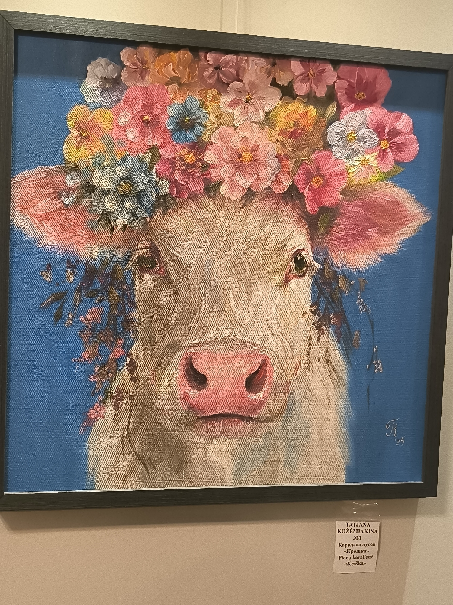 The image depicts a framed painting of a cow adorned with a floral crown. The cow has a light-colored coat and large, pink ears. The floral crown features a variety of colorful flowers, including pink, orange, yellow, and blue. The background of the painting is a solid blue. There is a signature and date in the bottom right corner of the painting. A small white label is attached below the painting, providing information in what appears to be Cyrillic and Latin scripts. The label includes the artist's name "TATJANA KOZEMIAKINA", and what seem to be a number "№1", a title in Cyrillic "Королева лугов", a title in quotation marks in Cyrillic "«Крошка»", and a title in Latin script "Pievų karalienė «Kroska»". The painting is displayed on a wall in what appears to be an indoor exhibition space. The wall is painted in a neutral color, and a portion of the floor is visible at the bottom of the image. The overall presentation suggests an art exhibition or gallery setting. The location of this exhibition is not specified in the image.