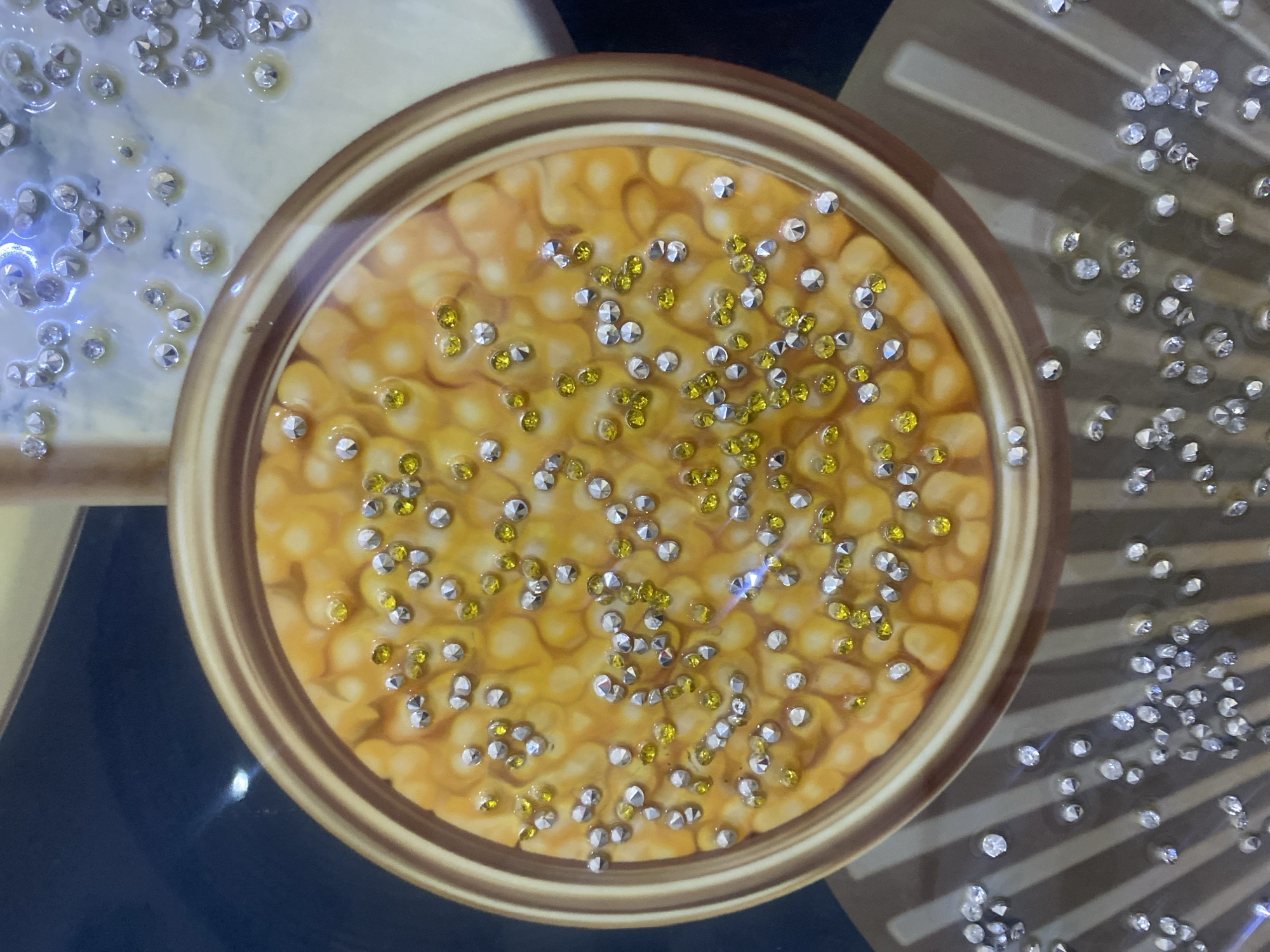 The image shows a close-up view of an ornate decorative piece. The central element is a circular inlay with a golden rim, filled with a textured orange-yellow material that resembles corn kernels. Scattered across this surface are numerous small, clear and yellow rhinestones, adding a sparkling effect. Surrounding this circular inlay, and extending downwards, is a striped pattern in shades of brown and beige. More scattered rhinestones are visible on this striped background, some appearing to be embedded in a glossy finish. The overall impression is of a highly decorative and possibly handmade item, with elements of sparkle and intricate design. The image does not depict any specific location in Giginyu, Nigeria, nor does it contain any people or actions.
