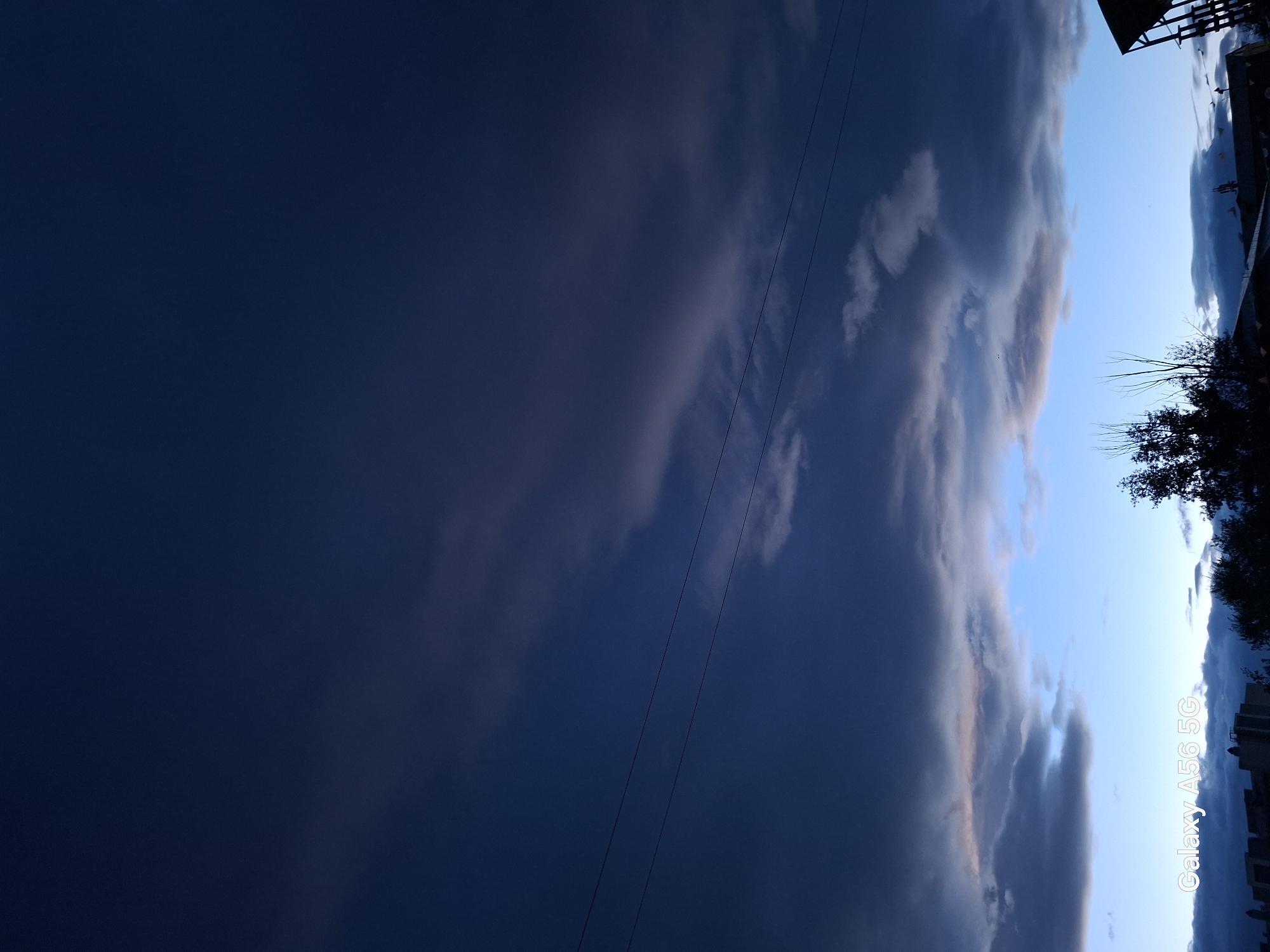 This is a low-angle photograph of a cloudy sky above the city of Chita, Russia. The sky is a deep, dark blue, with lighter patches of clouds interspersed throughout. Two thin power lines stretch horizontally across the upper portion of the frame. In the lower portion of the image, a sliver of lighter blue sky is visible, with dark silhouetted buildings and trees against it. The text "Galaxy A56 5G" is overlaid in white in the bottom left corner of the image.