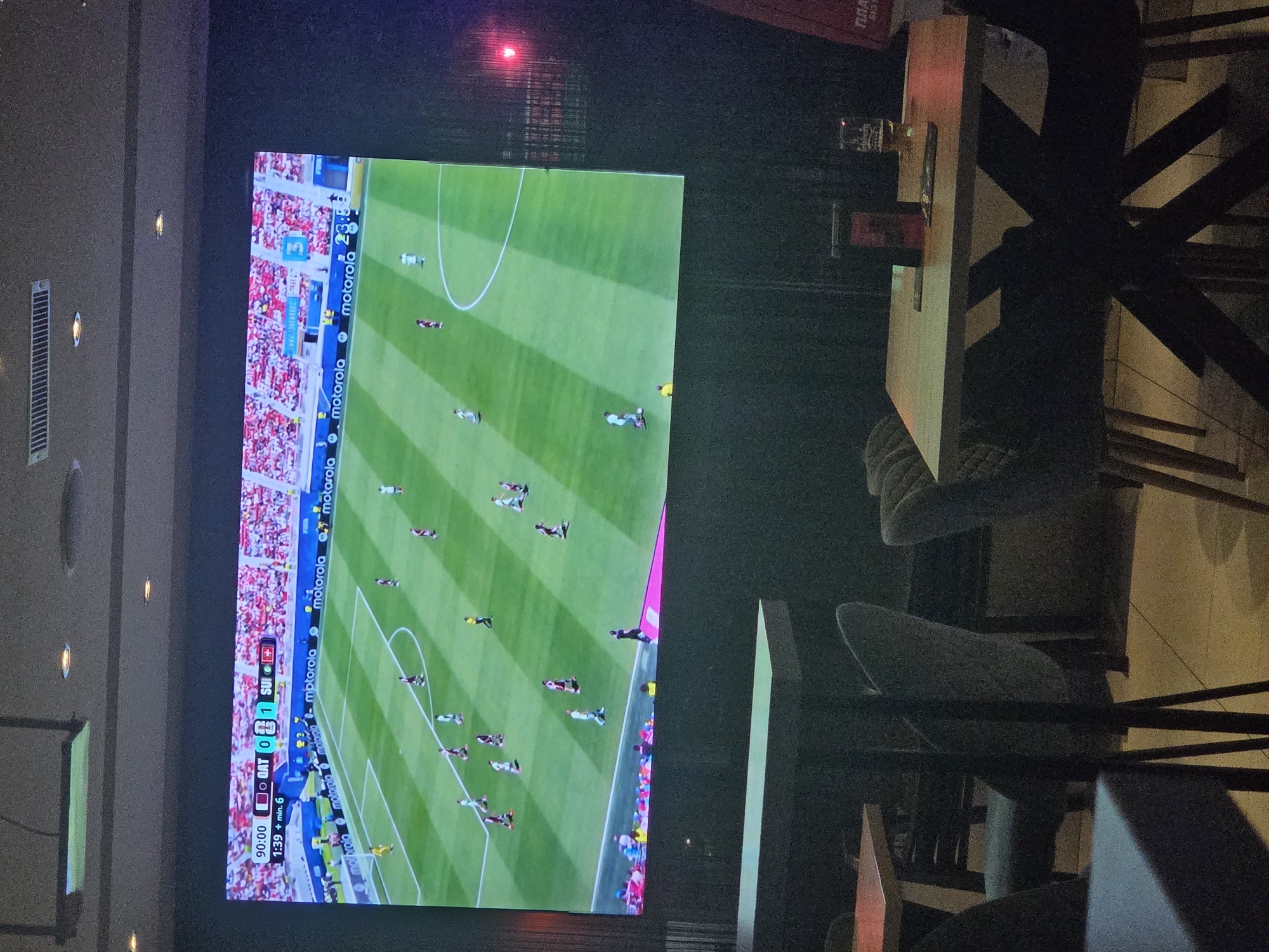 The image shows a large screen displaying a soccer match. The score on the screen indicates "QAT 0 - 1 SUI", with the time at "90:00 + min. 6". Several players in white and red uniforms are visible on the green field, with spectators in the background. The screen is positioned in a dark room, likely a bar or restaurant, as evidenced by the tables and chairs in the foreground. One table has a glass of beer and a menu. The overall lighting suggests an evening setting. The location of Targovishte, Bulgaria, is not discernible from the image itself, and any assertion of it being in Targovishte would require external information.