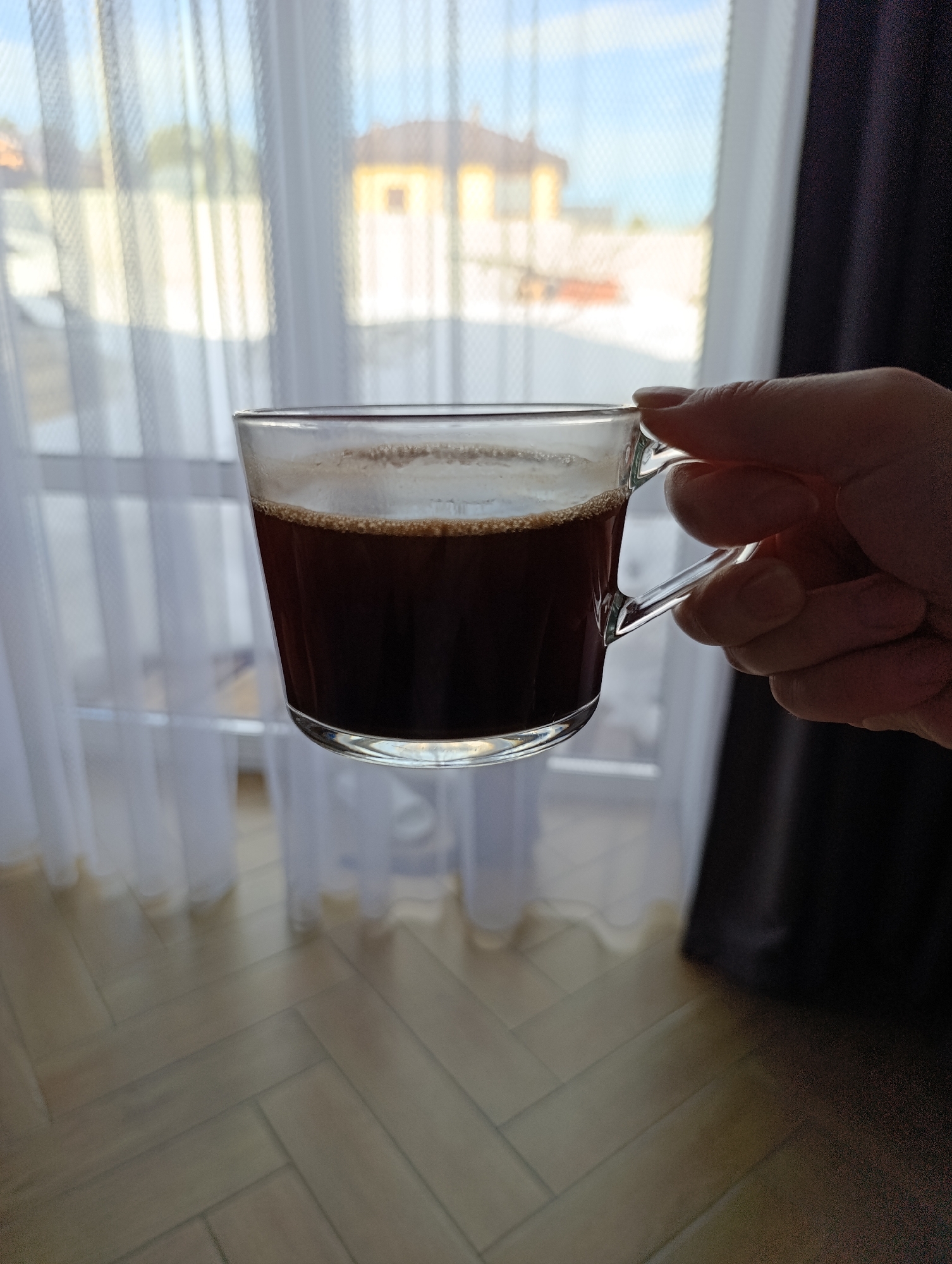An adult hand holds a clear glass mug containing dark brown liquid topped with light brown foam. The mug is centered in the frame, held by its clear handle. The background consists of an interior window with sheer white curtains and a dark curtain on the right. Through the window, an out-of-focus exterior scene is visible, depicting a building with a yellow facade and a dark roof, light-colored ground, and a blue sky. The floor in the interior foreground features light-toned planks arranged in a herringbone pattern. The scene is located in the city of Chaikovsky, Russia.