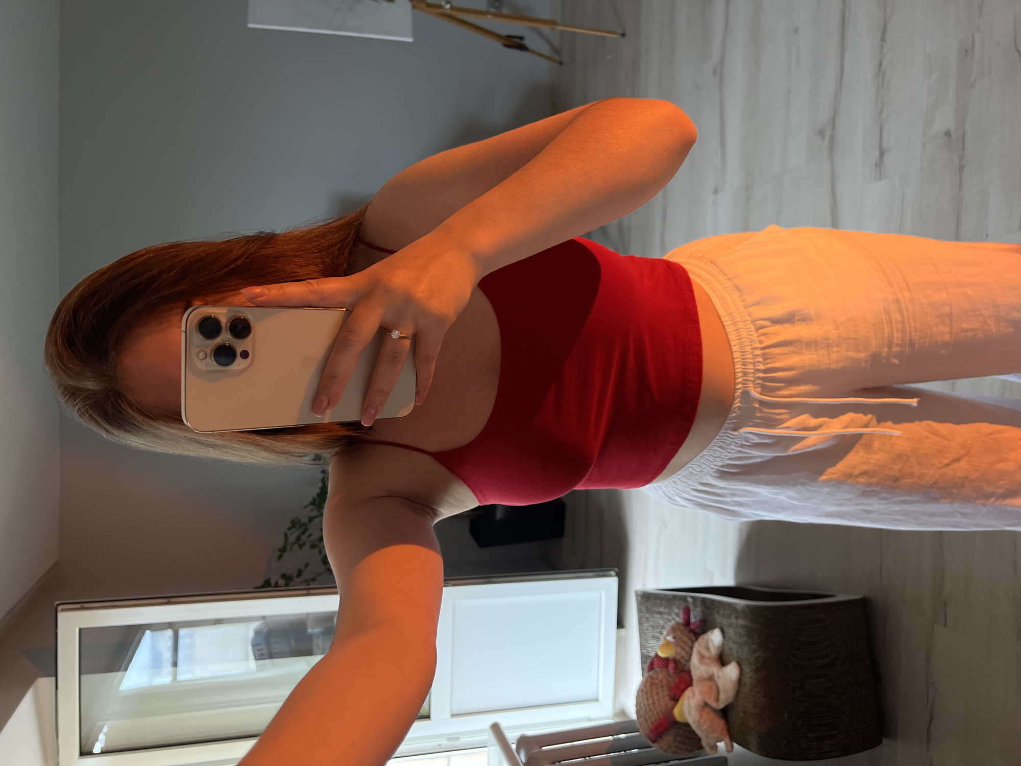 An individual is depicted in a mirror selfie, primarily showing their upper body, arms, and hair. They are wearing a red tank top or crop top paired with white, loose-fitting trousers that have a visible drawstring waist. A ring with a single clear stone is visible on the ring finger of their left hand, which holds a light-colored smartphone (an iPhone with a triple camera array) to capture the image. The person's fingernails are manicured with a French tip style.
The setting appears to be an indoor room, likely a home, with light grey walls and a light-colored wooden floor. Golden, warm light bathes the scene, suggesting either early morning or late afternoon sunlight streaming in. In the background, a light-colored wooden easel with a canvas is partially visible. A window with a white frame reflects light and what seems to be the exterior environment. Below the window, there's a decorative wooden box or planter adorned with two small, whimsical chicken figurines made of straw-like material. No discernible text is visible in the image. The activity taking place is a mirror selfie, obscuring the person's face. The location context is Ternopil, Ukraine.