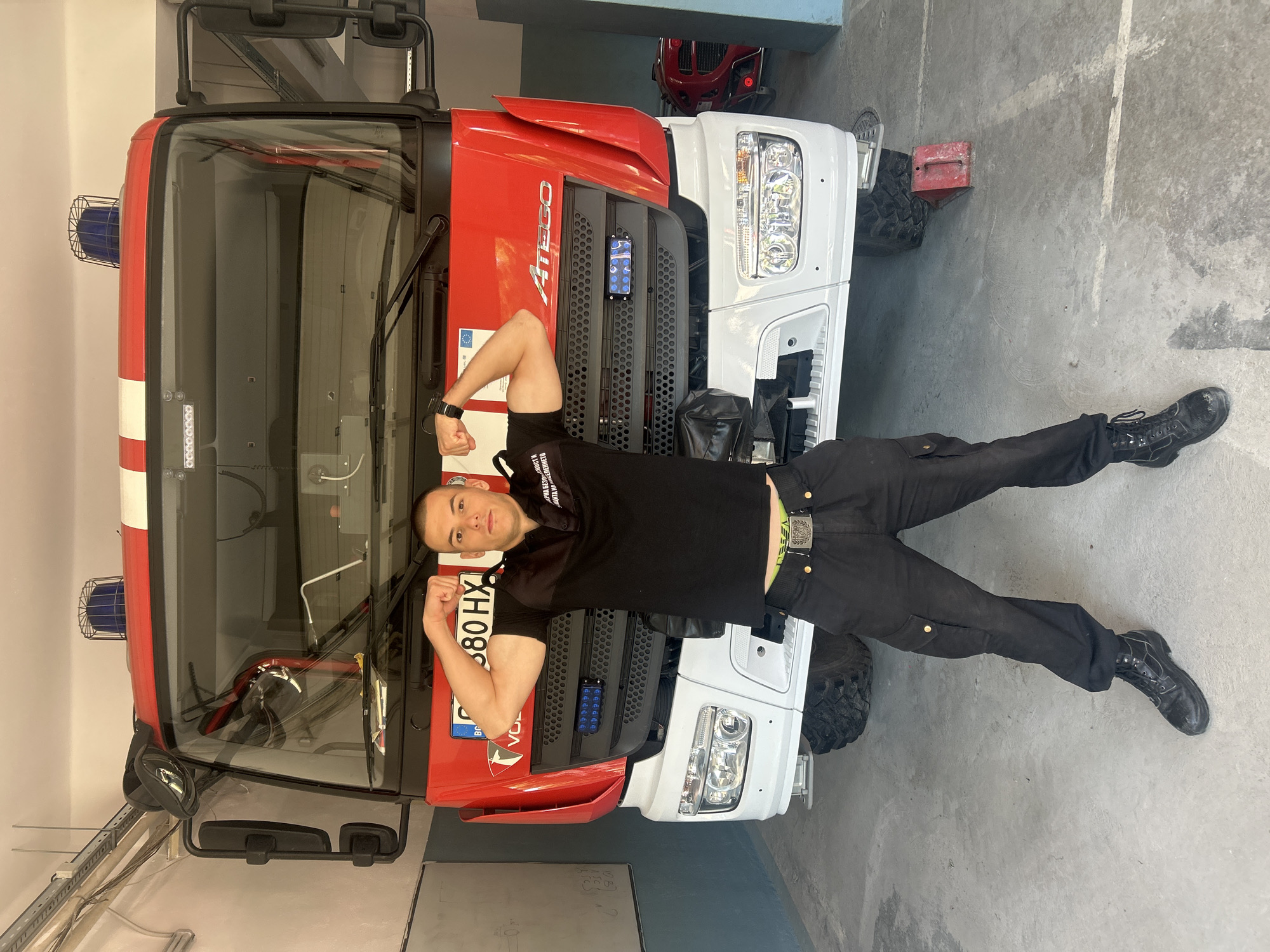 A man in a black shirt and pants is lying on a red and white fire truck. He is flexing his arms and looking at the camera. The fire truck has blue emergency lights on top and the words "ATEGO" and "VOL" on the front. A license plate reads "B 80 HX." Some text in Bulgarian can be seen below it on the red part of the truck. The truck is parked in what appears to be a garage or fire station. The setting implies that this could be in Bulgaria because the license plate is Bulgarian. Based on the details extracted from the license plate and the Bulgarian text on the vehicle, the general location of Sofia, Bulgaria can be inferred.