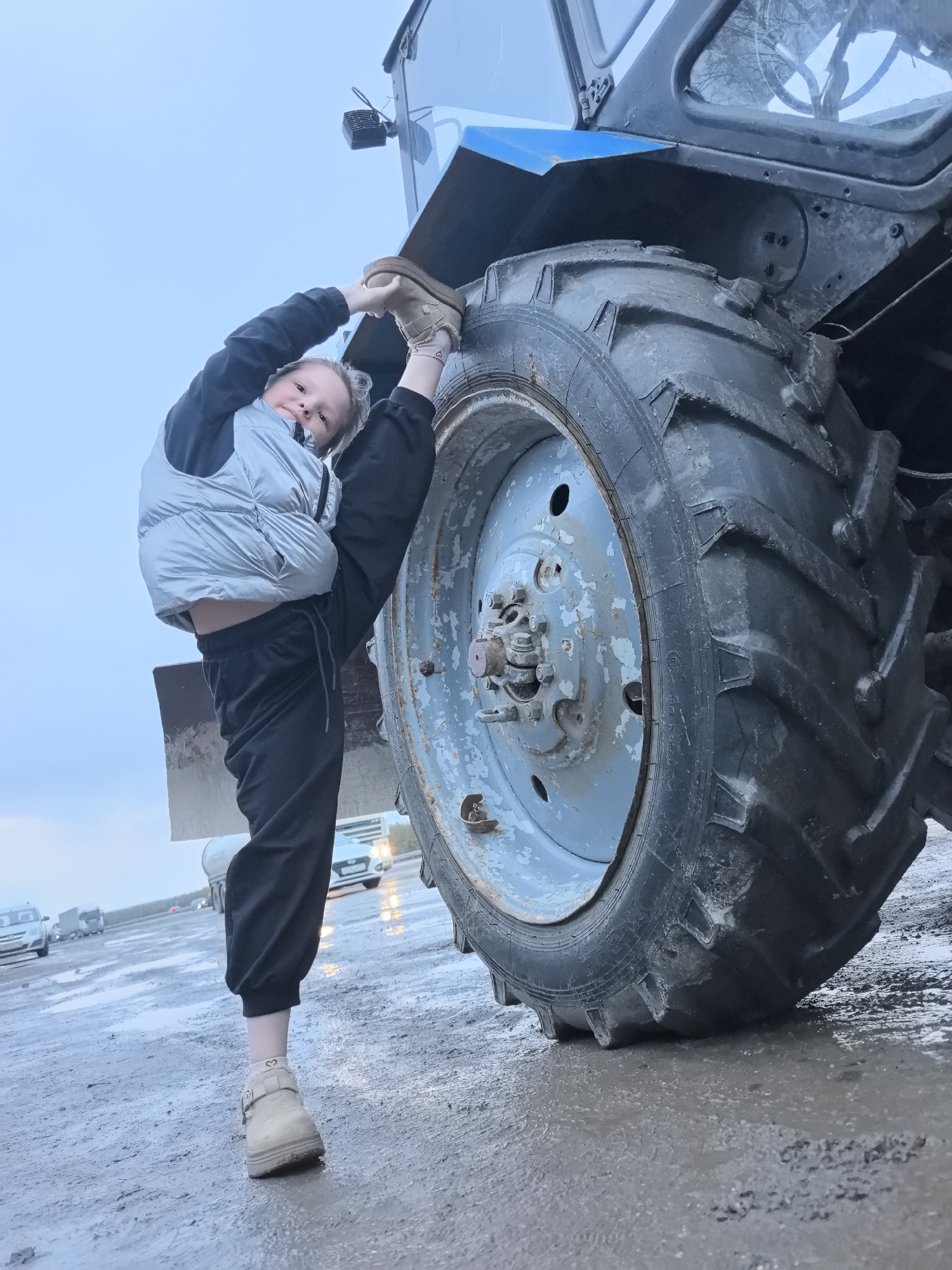 A young girl is performing a split next to a large tractor wheel. She is wearing a gray puffer vest, black sweatpants, and tan shoes. She is holding her other foot up to the tractor. The background shows a wet road and other vehicles. The location is Bol'shoi Mashlyak, Russia.