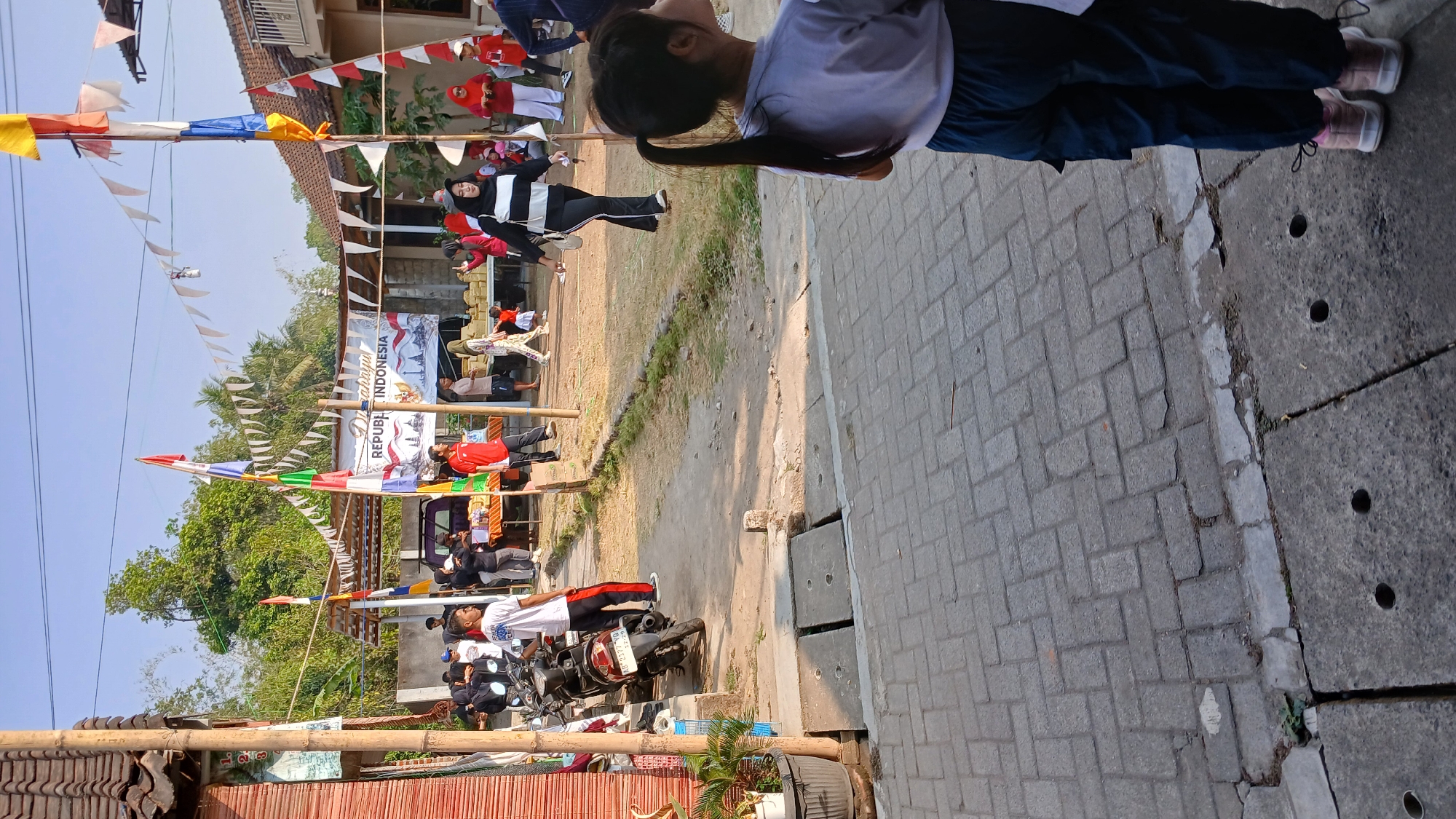 In Mendut, Indonesia, a scene unfolds with a festive atmosphere under a clear blue sky. The foreground features a young girl with dark hair in a ponytail, wearing a light purple shirt and dark blue pants, looking away from the camera. Her feet are clad in pink sneakers. To her left, a paved walkway is bordered by a concrete gutter containing drainage holes. Beyond the walkway, a dirt path leads to a gathering of people. Several individuals, including men and women, are present, some wearing traditional Indonesian clothing. Flags and pennants adorn the area, adding to the celebratory mood. In the background, a building with a red roof and wooden walls stands, alongside trees and other structures. A motorcycle is parked to the left, and tables laden with goods suggest a market or event. The overall scene depicts a community gathering, possibly a celebration or a local market, in Mendut, Indonesia.