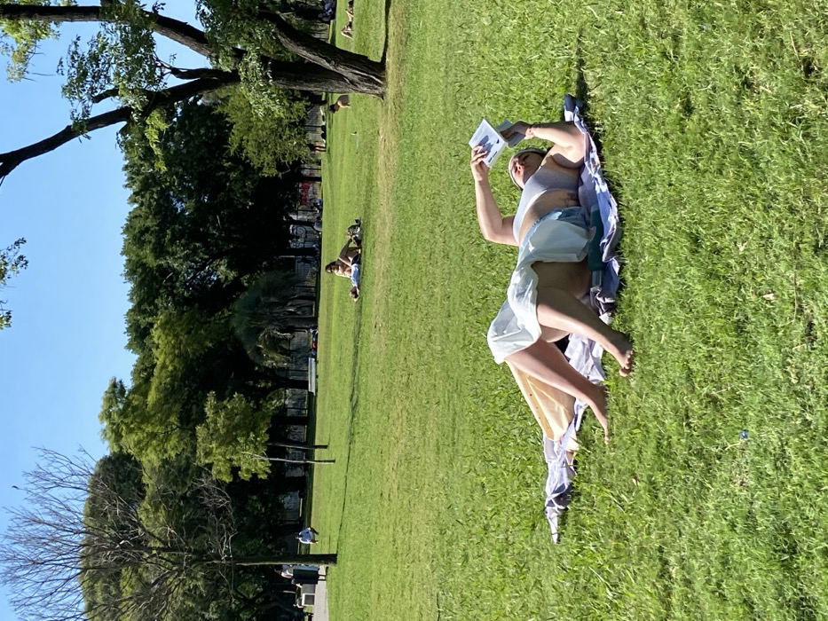 A woman in light clothing and a bikini top is lying on a blanket in a grassy park, reading a book. The sun is shining brightly, and the grass is green. There are other people relaxing on the grass in the distance, including a pair sunbathing in the middle. Trees and blue sky fill the background.