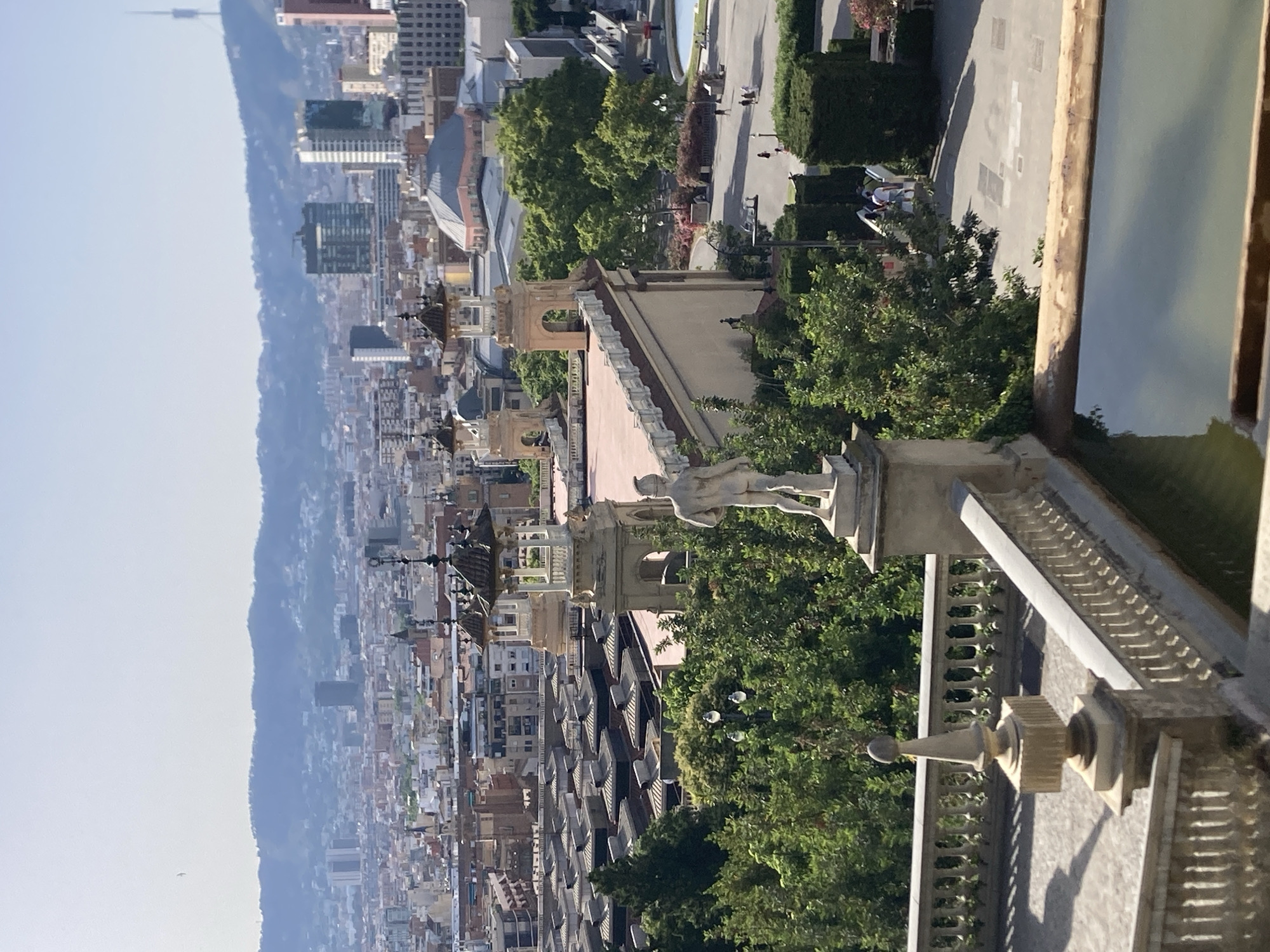This is a medium-shot, eye-level, exterior photograph of a city skyline. The scene is a panoramic view of Barcelona, Spain, taken from a high vantage point. In the foreground, a light-colored balustrade with decorative finials lines the edge of a terrace, with a pool of water visible in the lower right corner. A stone statue of a nude male figure stands on a pedestal behind the balustrade.
In the midground, the complex of buildings below features a distinct row of rooftop structures with dark, pitched roofs, possibly market stalls or a similar commercial area. Behind this, several ornate, open-sided pavilions with domed roofs rise above the trees. Lush green trees form a dense canopy in front of these structures, interspersed with the architecture of the city.
The background reveals the sprawling urban landscape of Barcelona, characterized by a dense collection of buildings of varying heights and architectural styles. Tall modern skyscrapers punctuate the cityscape, and in the distance, a hazy mountain range forms the horizon under a bright, clear sky. A distinctive tower, likely a broadcast or observation tower, is visible on one of the distant hills. The overall impression is a sunny day, with the light casting shadows across the terrace in the foreground. Several people are visible walking on pathways and in open areas in the midground, indicating public access and activity within the area.