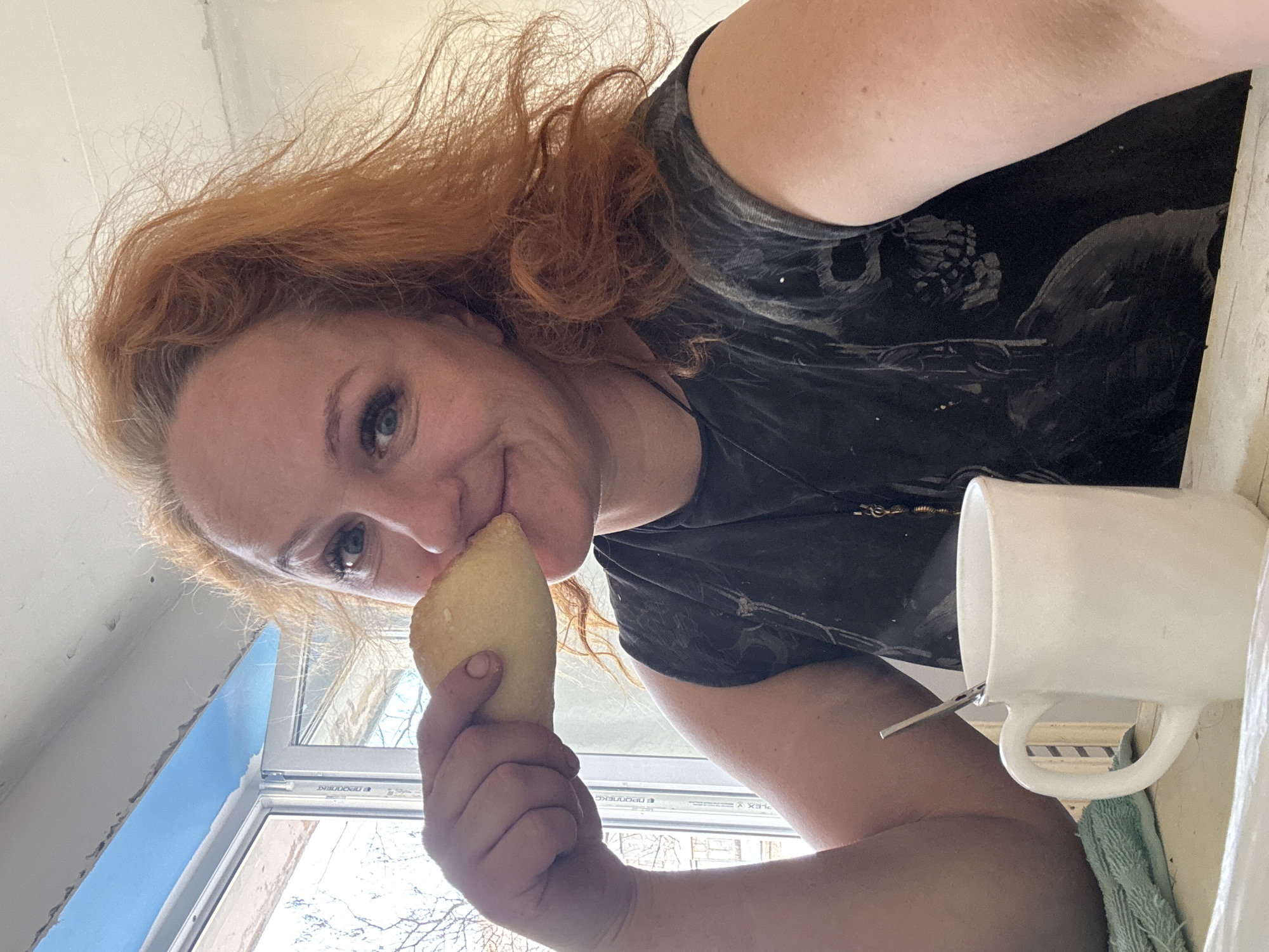 A woman with red hair and blue eyes smiles playfully at the camera in a rotated, close-up shot. She holds a golden-brown, teardrop-shaped pastry to her nose, as if smelling it. She wears a dark t-shirt with a faint, possibly skull-themed, pattern. To her right, a white ceramic mug with a spoon or stirring stick rests on a surface, beside a green cloth. The background reveals an indoor setting with natural light streaming through a window. Bare tree branches are visible outside the window, suggesting a colder season like winter or early spring. A section of a light blue wall is seen through the window opening. Visible text on the window frame includes "ПРОПОНЕС", "PLEX A", and parts of the "DECEUNINCK" brand logo, indicating PVC window profiles. The woman's expression is happy and relaxed. The scene appears to be set indoors, likely in a home in Chaikovsky, Russia, during daytime.