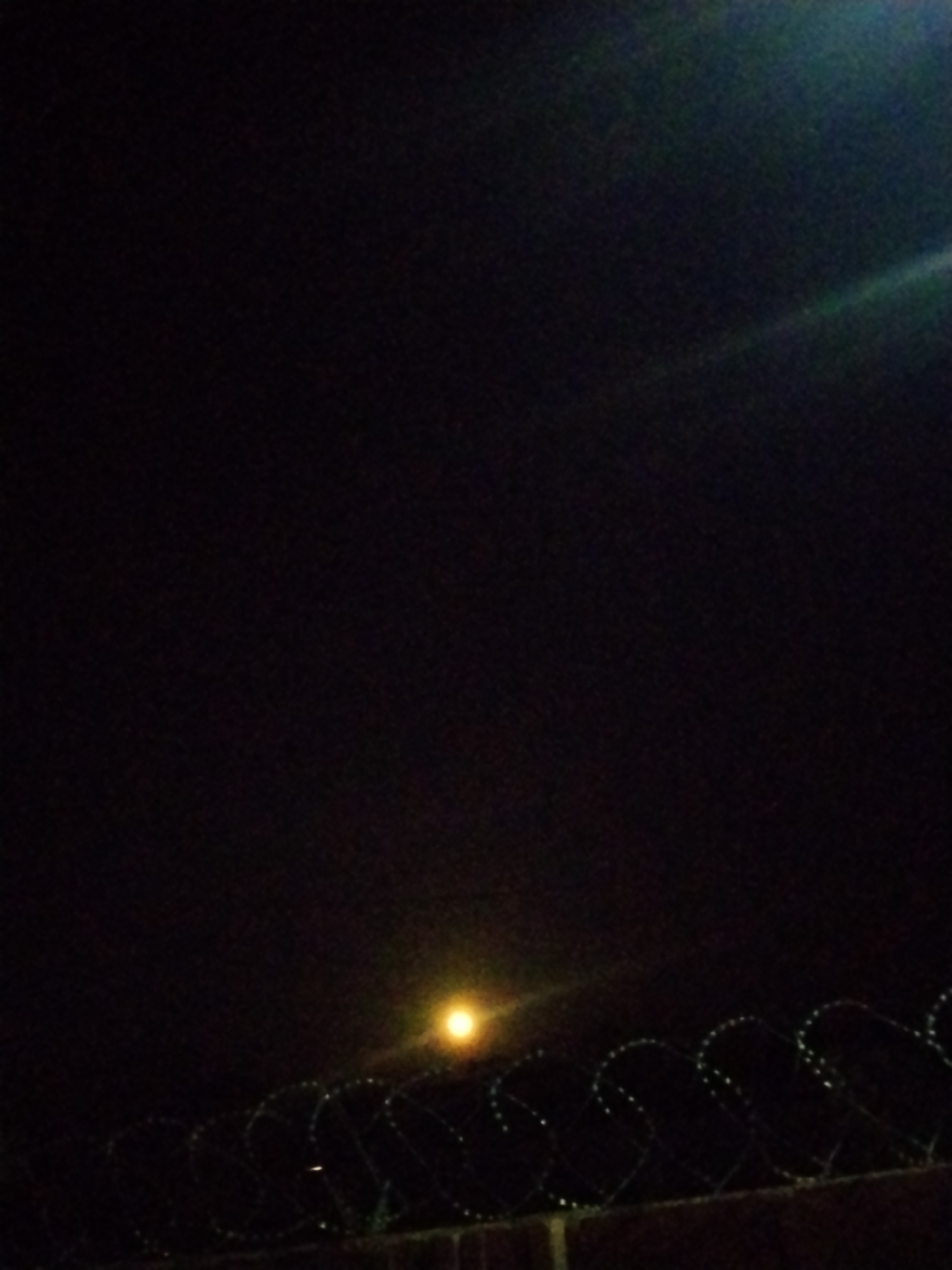 The image is a low-light, slightly grainy photograph taken at night in Mangu Town, Nigeria. The scene is dominated by a dark sky with a faint, hazy illumination from an unseen light source, possibly the moon or a distant streetlamp, positioned in the lower middle portion of the frame. A bright, circular light source, appearing yellow-orange, is visible in the center of the image, casting a soft glow. In the foreground, a coiled barbed wire fence stretches horizontally across the bottom third of the frame. The fence appears to be mounted on a wall, likely made of brick, though the detail is obscured by darkness. The barbed wire itself is faintly illuminated, highlighting its sharp, circular patterns. The overall impression is one of a secured perimeter or boundary, viewed under nocturnal conditions.