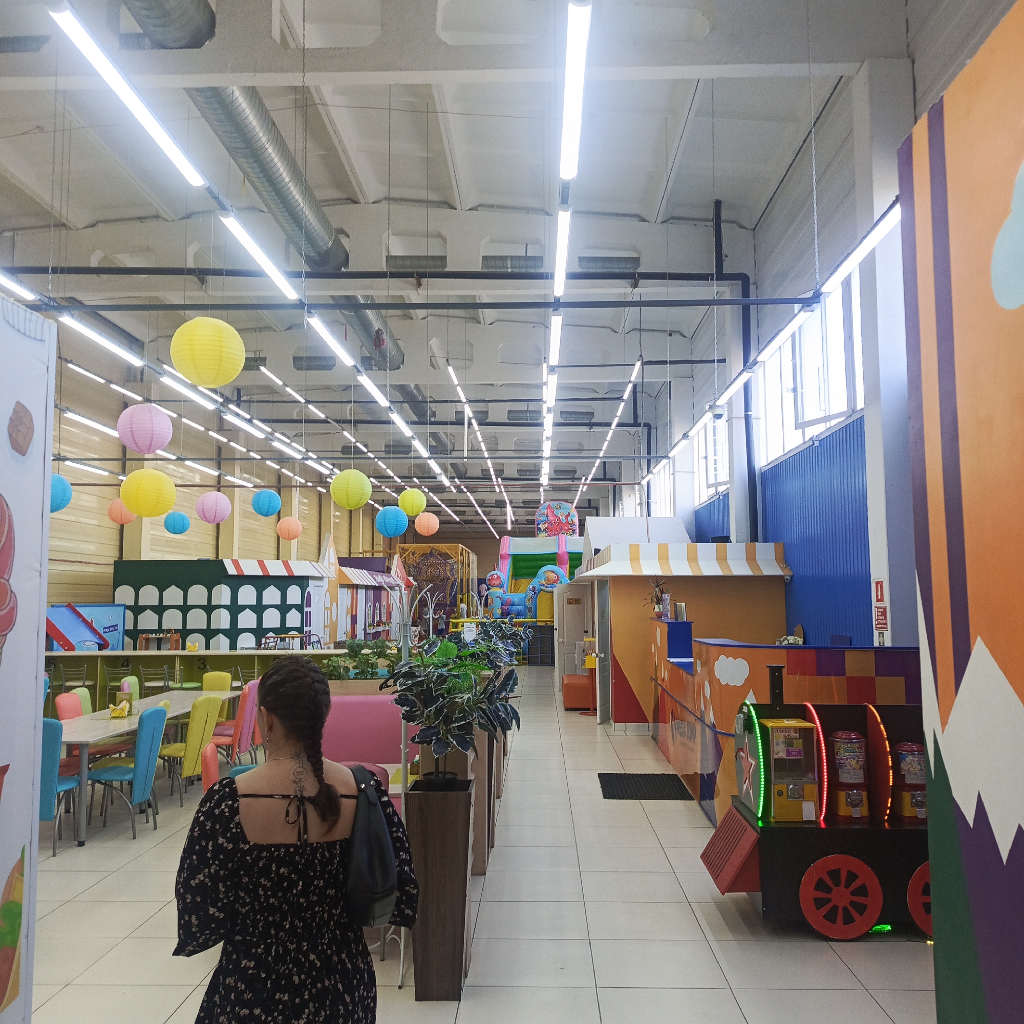 The image shows the interior of a children's entertainment center in Chaikovsky, Russia. The space is brightly lit with fluorescent lights suspended from a high, industrial-style ceiling. Colored paper lanterns hang in the distance. A woman with a braid and floral dress stands with her back to the camera. There is a play area including small colorful playhouses, a bouncy castle, tables and chairs for dining, and some plant-filled planters lining the central walkway. At the far end, windows provide natural light. To the right, is a toy train and a colorful reception desk.