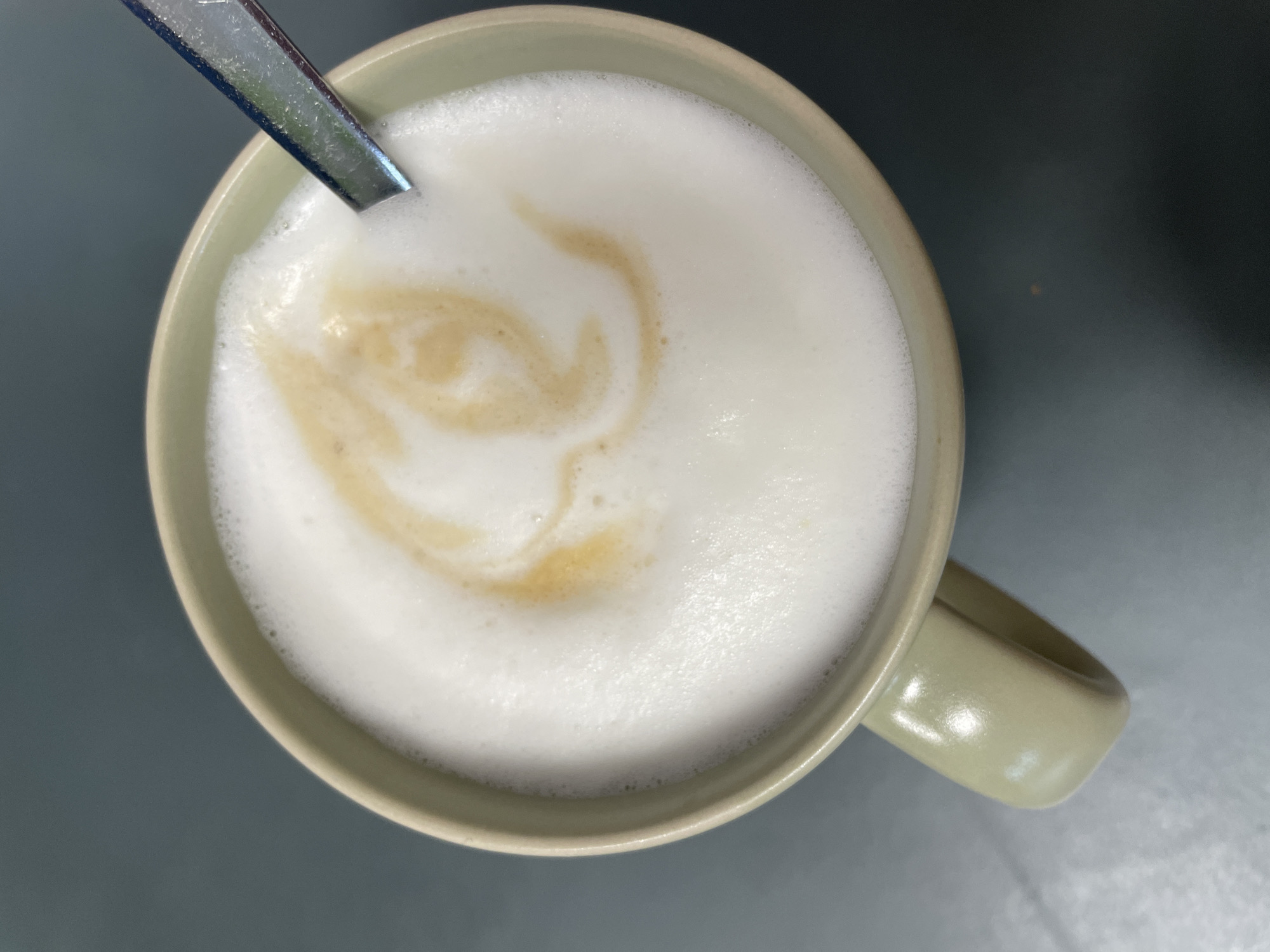 An overhead, close-up shot shows a tan mug filled with a frothy, white beverage, likely coffee or latte, with a light brown swirl pattern on top. A silver spoon rests in the drink, with its handle visible and protruding from the upper right of the mug. The mug's handle is also visible on the left side of the frame, and the mug itself is a pale tan color. The beverage is topped with a thick layer of foam. The background is a dark gray, smooth surface, possibly a table or countertop, with a slight sheen indicating it might be reflective. There is no indication of the location in Morsbach, Germany.