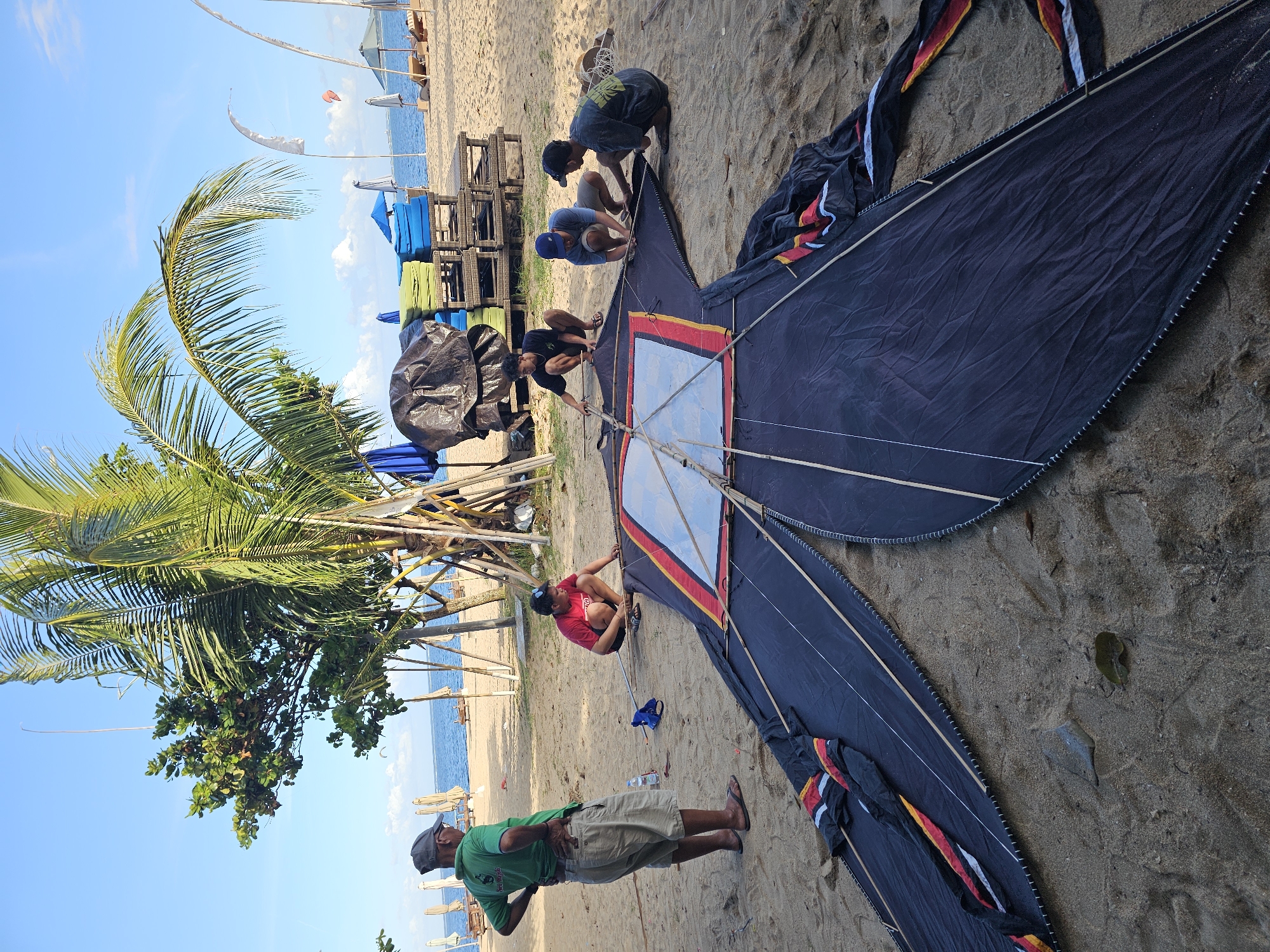 The image depicts a coastal scene in Denpasar, Indonesia. The location is on a sandy beach, evidenced by the presence of sand and a palm tree on the left. The ocean is visible in the background, and several beach umbrellas are positioned further down the coastline.
The primary subject is a large, assembled kite with a dark blue body, a square clear panel in the center, and colored accents of red, yellow, and white along the edges. A group of people are interacting with the kite. Three are crouched around the center section, possibly working on its assembly or repair. Another person in a red shirt is kneeling near the center of the kite, likely assisting. One man in a green shirt stands at the base of the kite.
Other objects of note include stacked chairs covered by a dark blue tarp, and beach umbrellas.