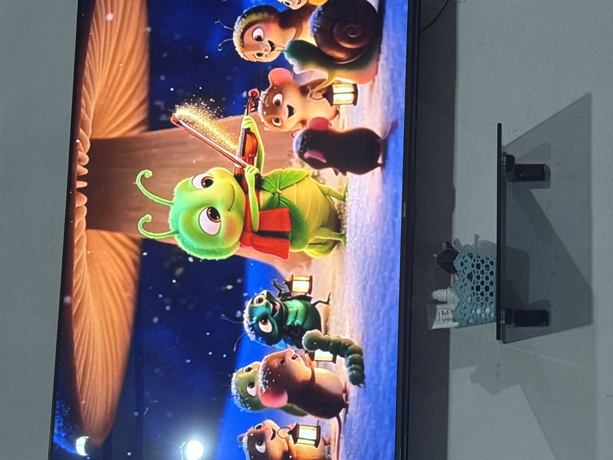 In the city of Chardara, Uzbekistan, a television screen displays an animated scene featuring anthropomorphic insects and small mammals gathered in a snowy, nighttime environment. A large, glowing mushroom serves as a backdrop. The central character, a green caterpillar wearing a red scarf, plays a violin, emitting golden sparks. Surrounding the caterpillar are several smaller creatures, including mice, a beetle, a snail, and a worm, each holding a lantern. The overall atmosphere is festive and magical, with snow falling gently and soft light emanating from the mushroom and lanterns. The television is mounted on a wall, and below it, a black shelf holds a light blue woven basket containing toiletries. Shadows are cast on the wall behind the shelf.