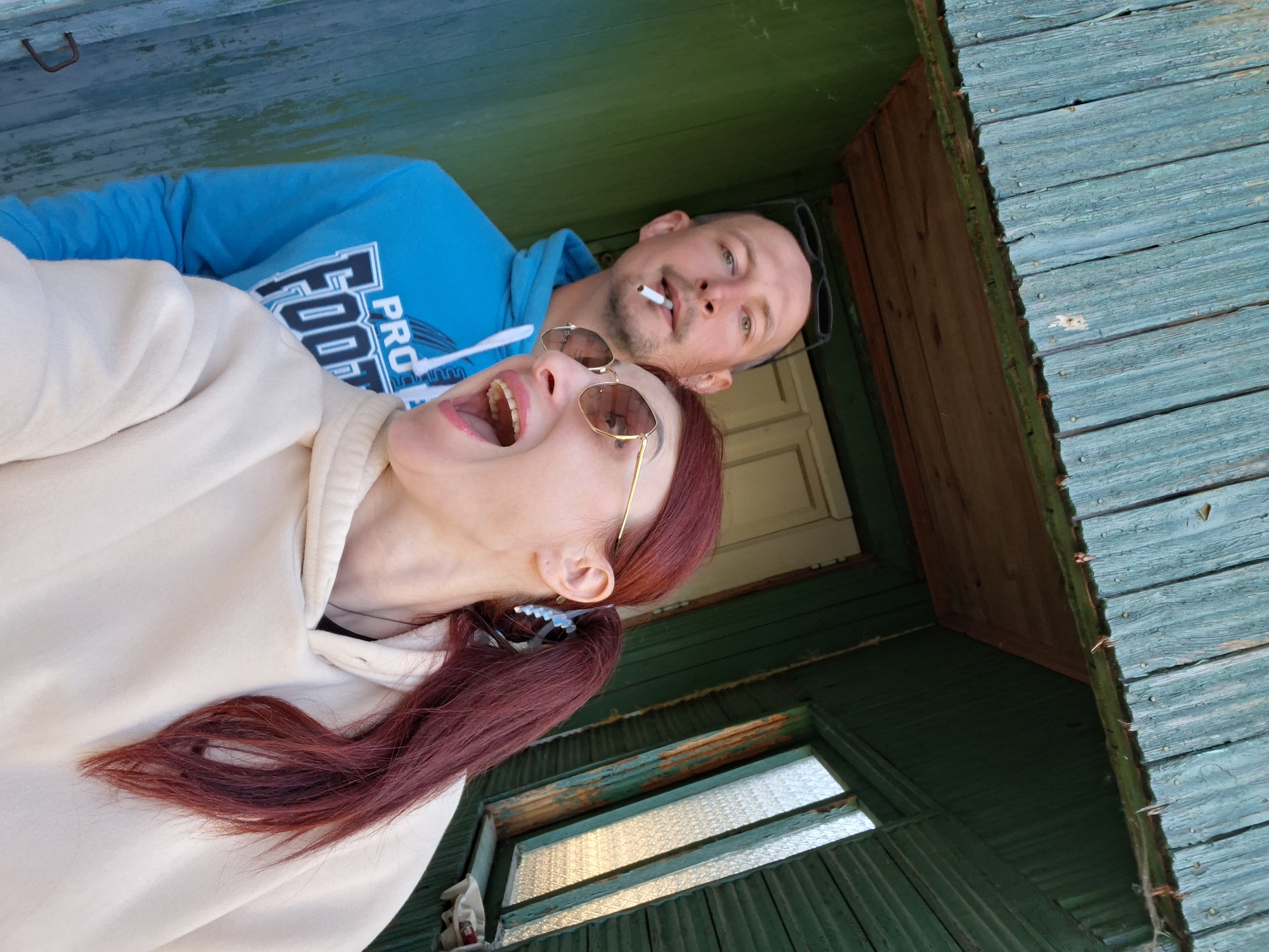 Two people are visible in the image, presumably a couple, against the backdrop of a weathered building exterior. A woman with auburn hair, styled in a low ponytail secured with a blue hair clip, wears a white hoodie and gold-framed sunglasses. Her mouth is open wide. Behind her is a man wearing a blue hoodie with "FOOTBALL" printed on the chest. He has a cigarette in his mouth and his black-framed sunglasses are resting on his head. The building's exterior is painted in shades of green and blue with chipped paint. A small window is visible in the lower portion of the frame. The location may be Забалоцкі сельскі Савет, Belarus.