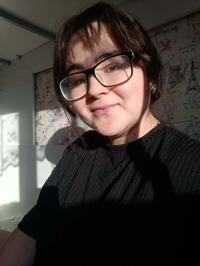 A female individual is depicted in a medium close-up portrait, located indoors within what is understood to be the city of Noviy, Russia. She has dark brown hair with bangs, wears black-framed glasses, and is looking directly at the camera with a subtle smile. She is dressed in a black, short-sleeved top with a vertical ribbed texture. A small, light-colored earring is visible on her right earlobe. The scene is illuminated by strong natural light entering from the left, creating a high-contrast effect. Direct sunlight illuminates the left side of her face and hair, while the right side of her face, neck, and torso, along with a significant portion of her clothing, are enveloped in deep shadow. The background consists of a wall covered in wallpaper featuring a pattern of pink and purple floral elements alongside sketches of Parisian landmarks, notably the Eiffel Tower, and text including "Mon Amour." Shadows consistent with window frames are cast upon the wallpaper to the left of the subject. A bright, unidentifiable white structure is visible along the upper left ceiling line.
