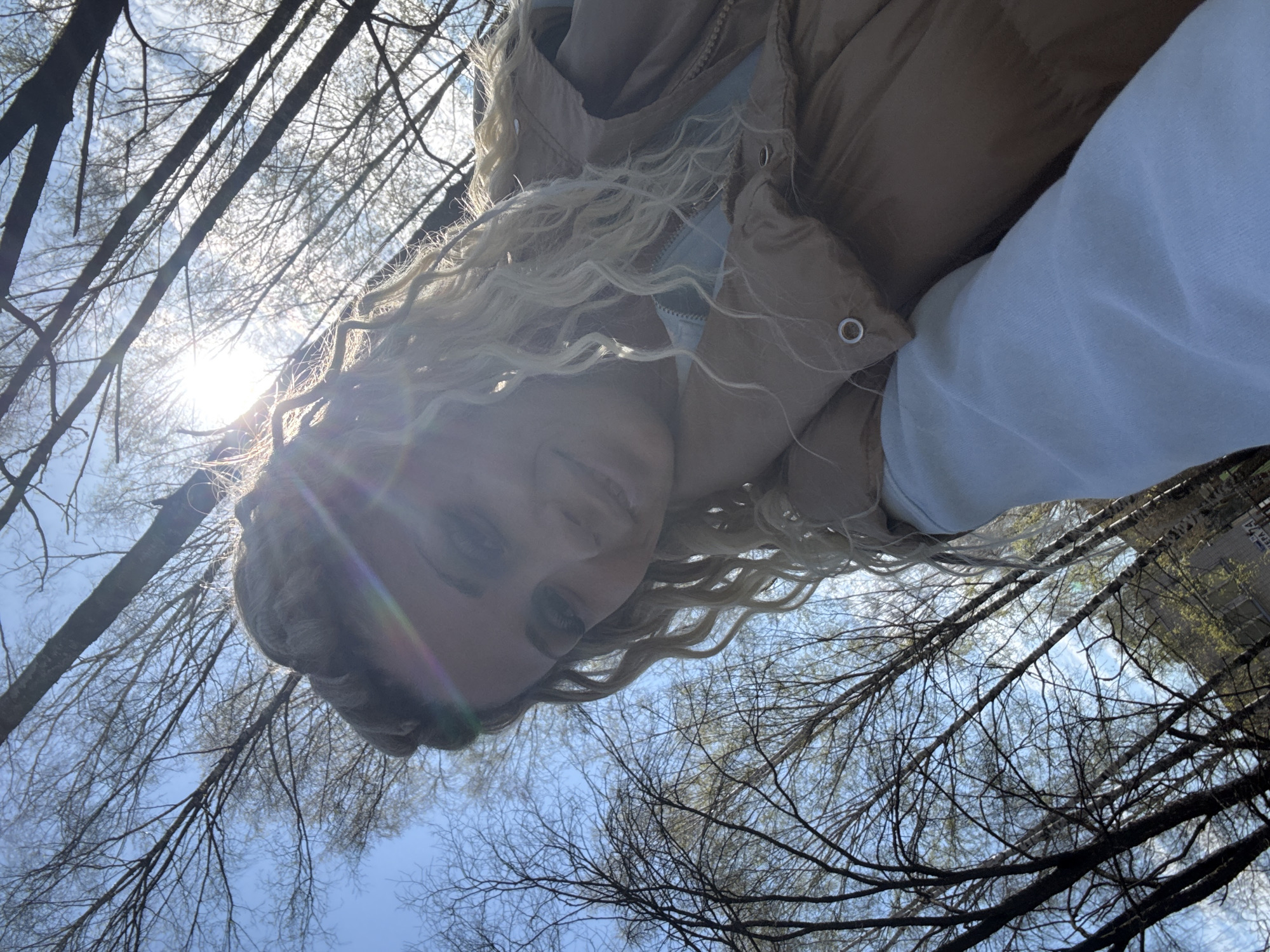 The image is an upward-facing shot of a woman, appearing to be in her 20s, standing outside. She has blonde, curly hair, and is wearing a light brown puffer jacket over a light-colored long-sleeve shirt. The sun is positioned directly behind her, causing a lens flare effect and making her face somewhat obscured. Above and around her are bare tree branches against a blue sky. The location appears to be a park or wooded area. Rusanovo, Russia is mentioned as the city where this image was taken.
