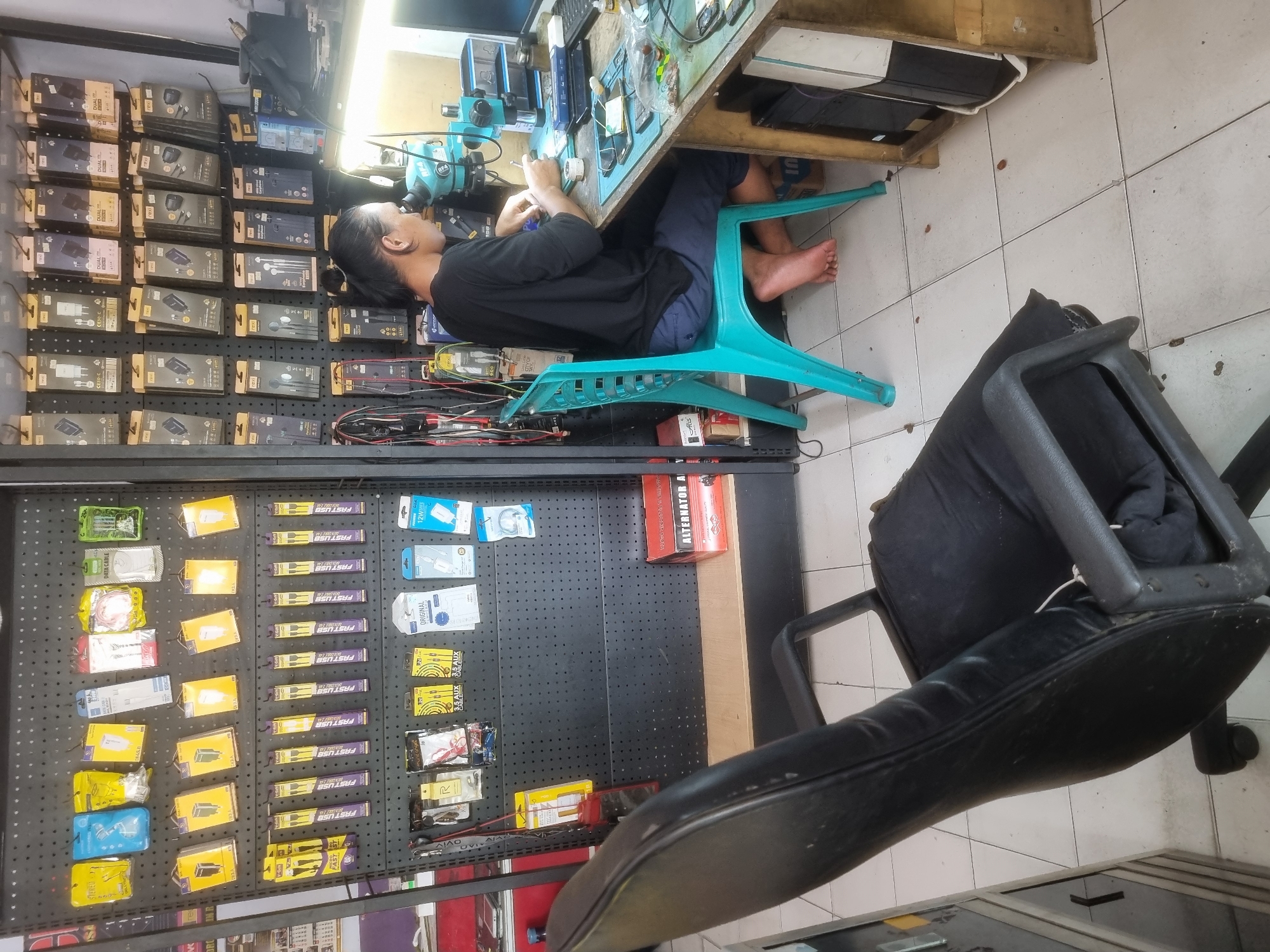 The image depicts a small electronics repair shop, possibly located in Probolinggo, Indonesia. A man is seated at a workbench, appearing to be engaged in intricate repair work on electronic devices, using a microscope for magnification. The workbench is cluttered with tools, electronic components, and various mobile phones. Shelves behind the workbench are filled with packaged electronic accessories, such as chargers, cables, and earphones. A black office chair is positioned in the foreground, partially obscuring the view of the lower half of the scene. The shop appears to have a utilitarian, organized display of goods, typical of small businesses in the region. The overall impression is one of a working environment focused on repairing and selling mobile phone accessories.